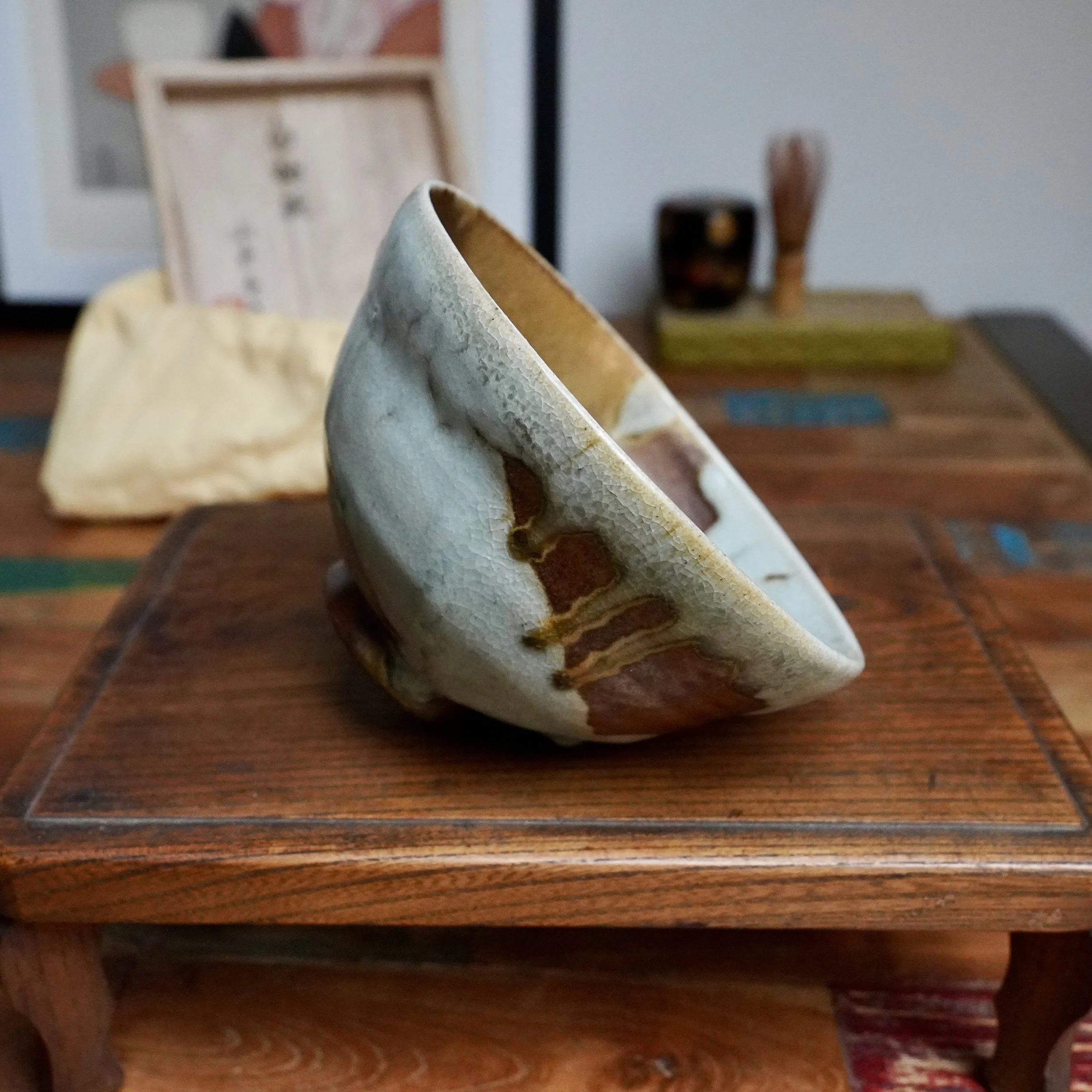 Angled chawan with crackled blue-grey glaze flowing into rust-brown, featuring ochre streaks and an unglazed red-brown foot, on a small wooden stand.