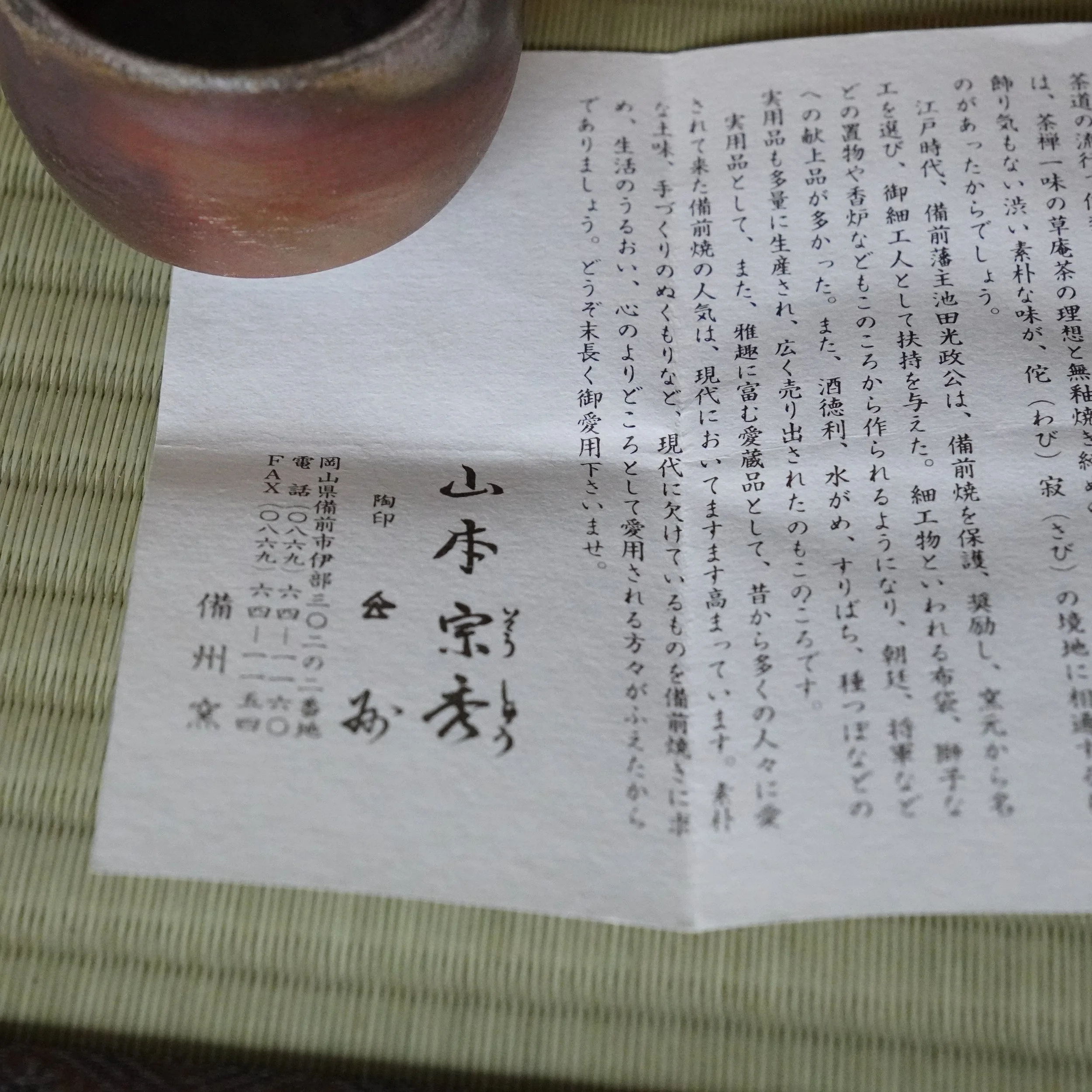Bizen-yaki guinomi with reddish hidasuki marks and an uneven rim, partially visible on a woven mat. A paper slip with Yamamoto Sōshū's calligraphic signature and Japanese text rests beside it.
