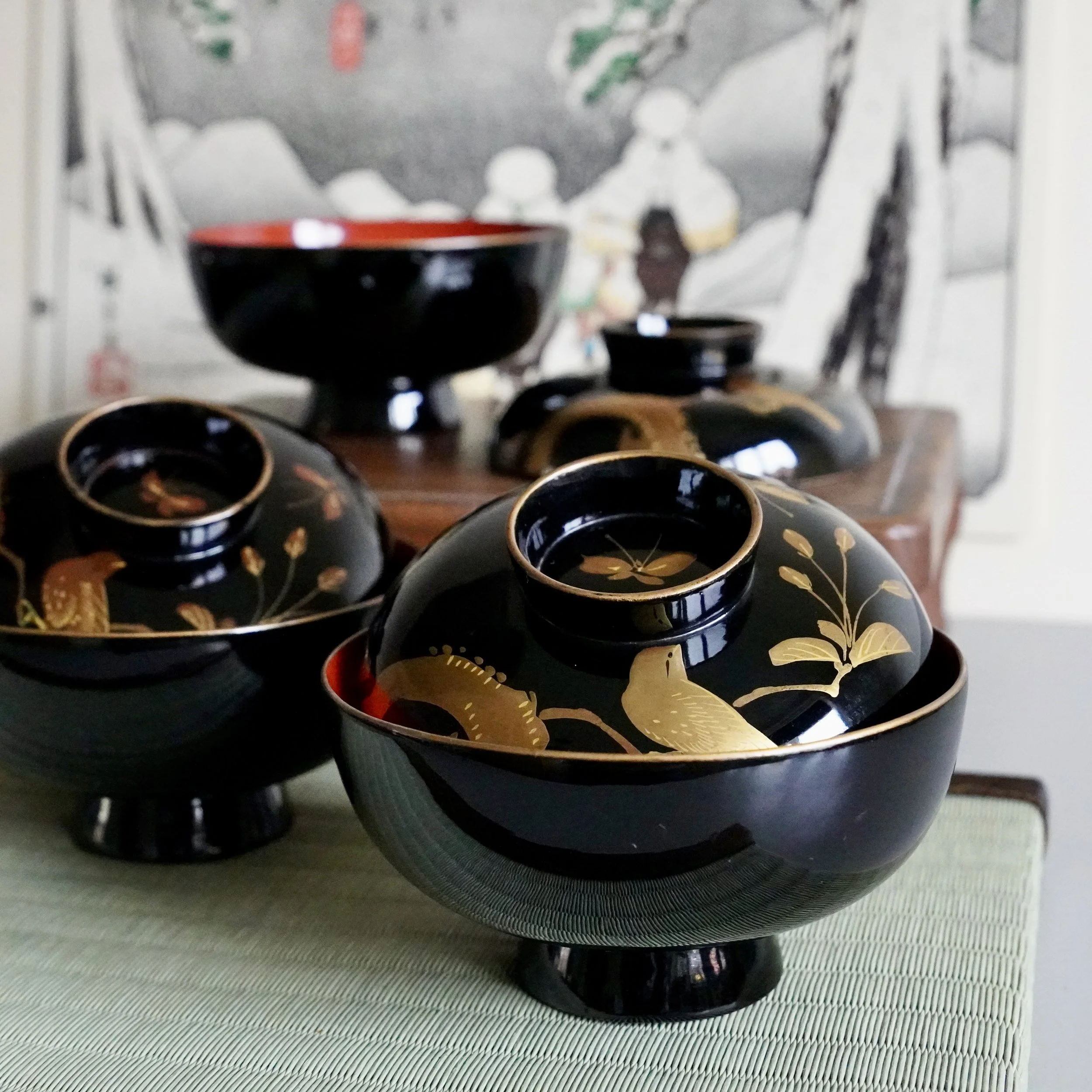 A set of black and gold urushi lacquer vintage antique bowls with floral designs, in front of a blurred background Japanese ukiyoe print