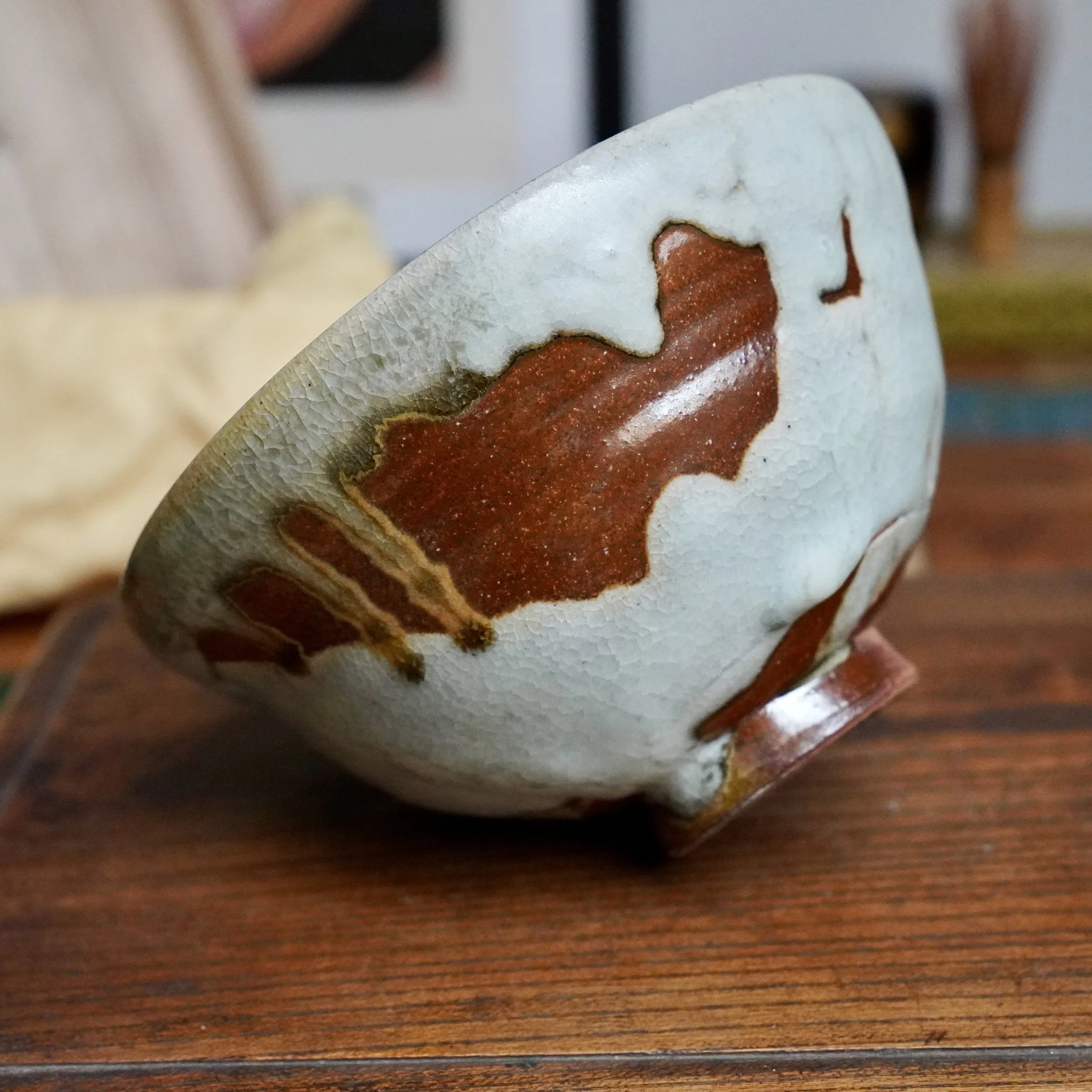 Side view of a chawan with a crackled pale blue-grey glaze flowing into a granular rust-brown, on a wood surface.