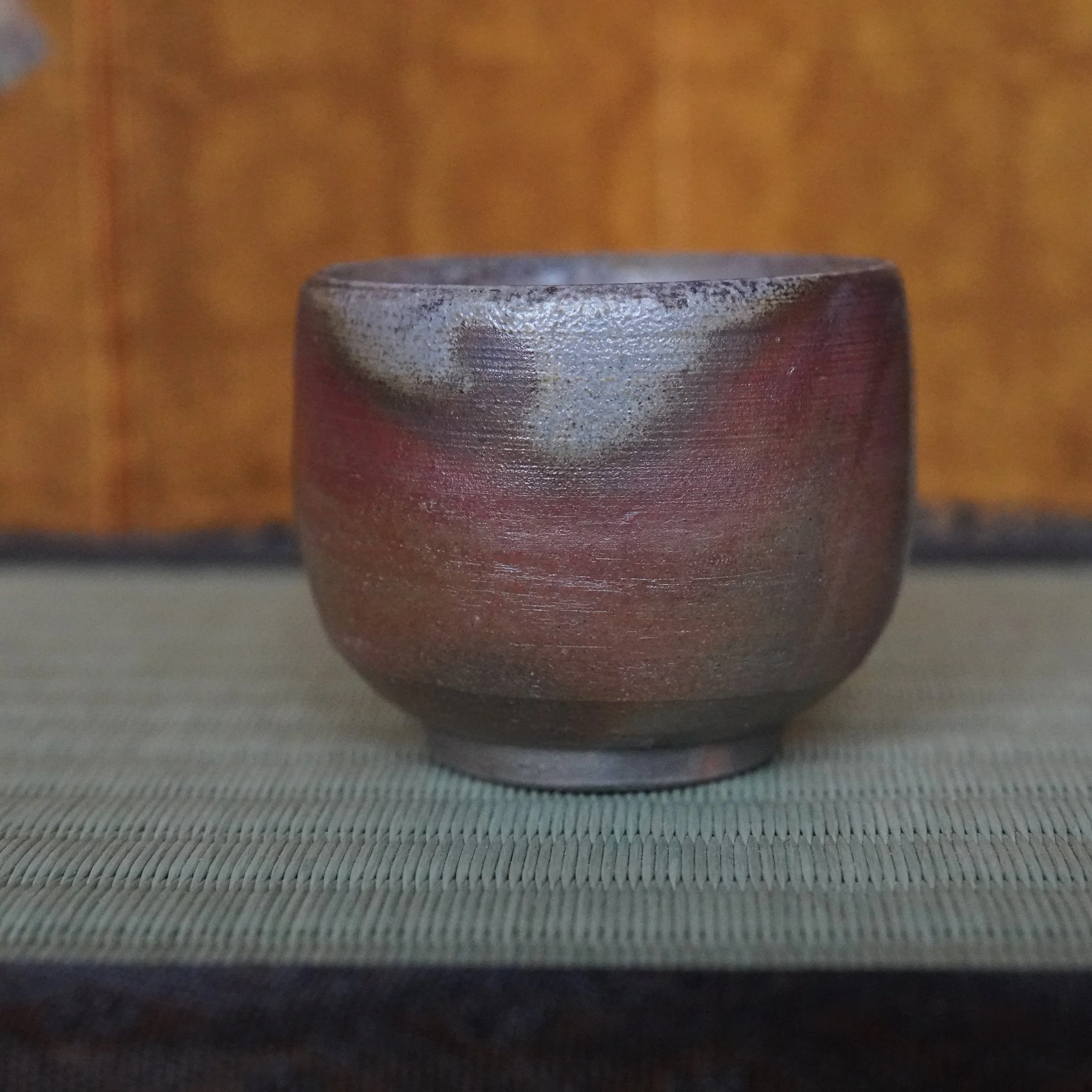 Yamamoto Sōshū Bizen-yaki guinomi with hidasuki flame marks in scarlet and grey on an ochre body, resting on a woven mat.