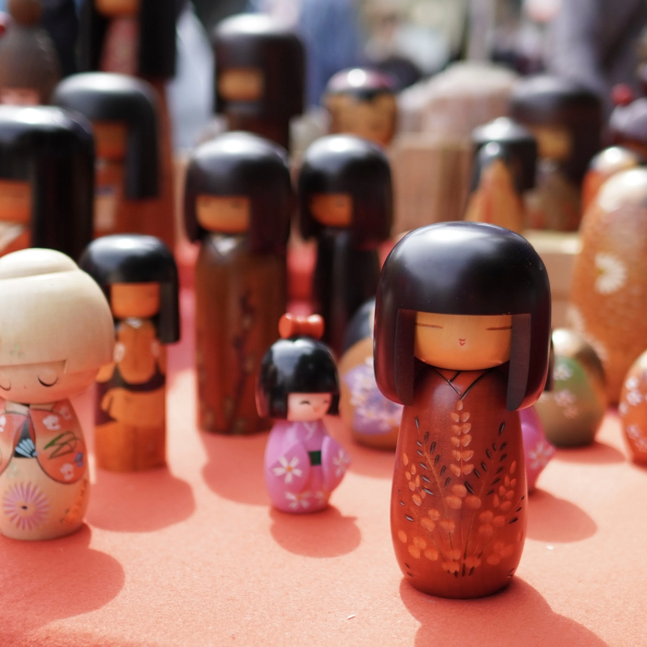 A group of vintage wooden kokeshi dolls in various sizes and styles, featuring hand-painted floral patterns and traditional expressions, displayed on a bright red surface at a market.