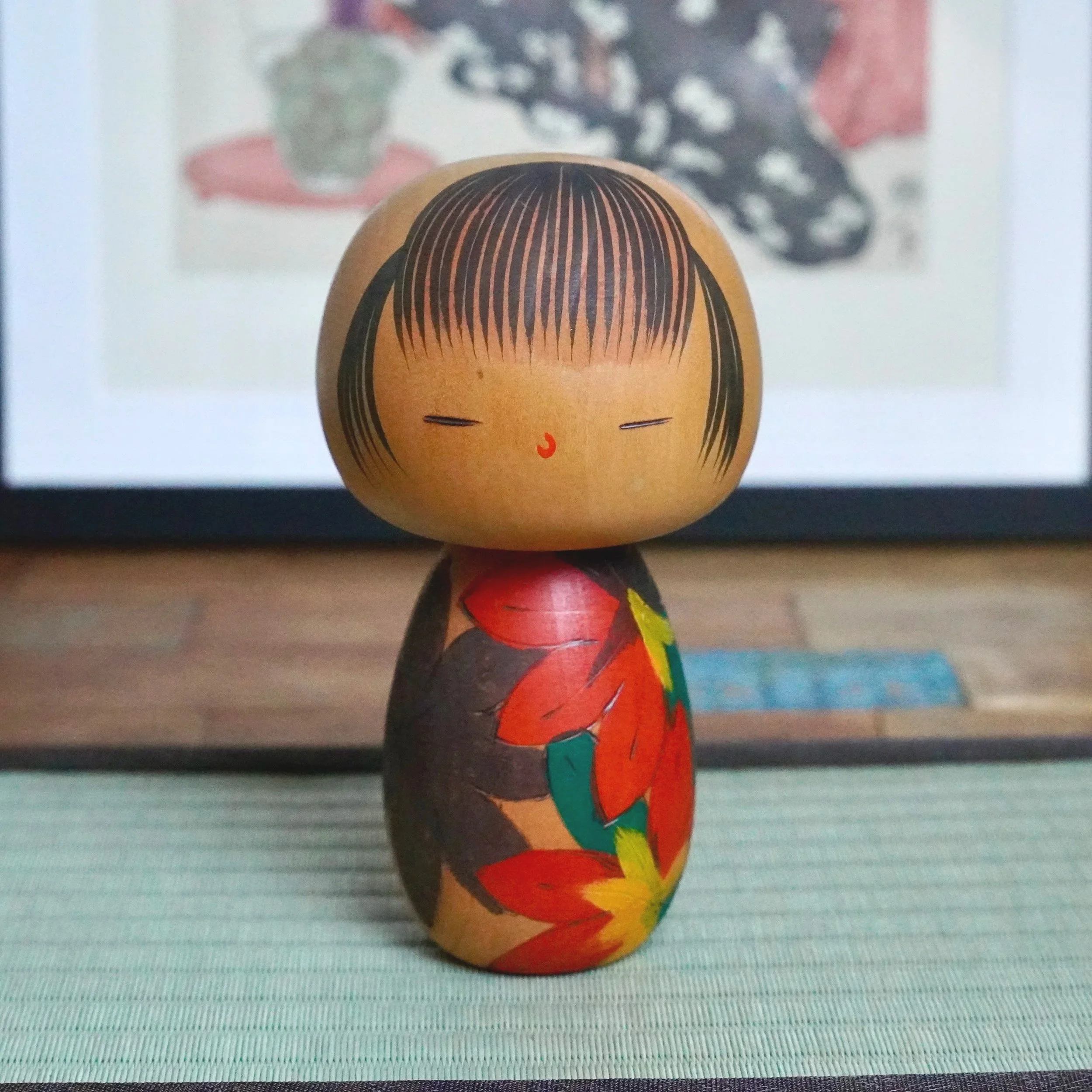 Kokeshi doll with a light wood head, closed eyes, red nose, and black hair. Its body features carved red, dark brown, green, and yellow autumn leaves. Stands on a green mat.