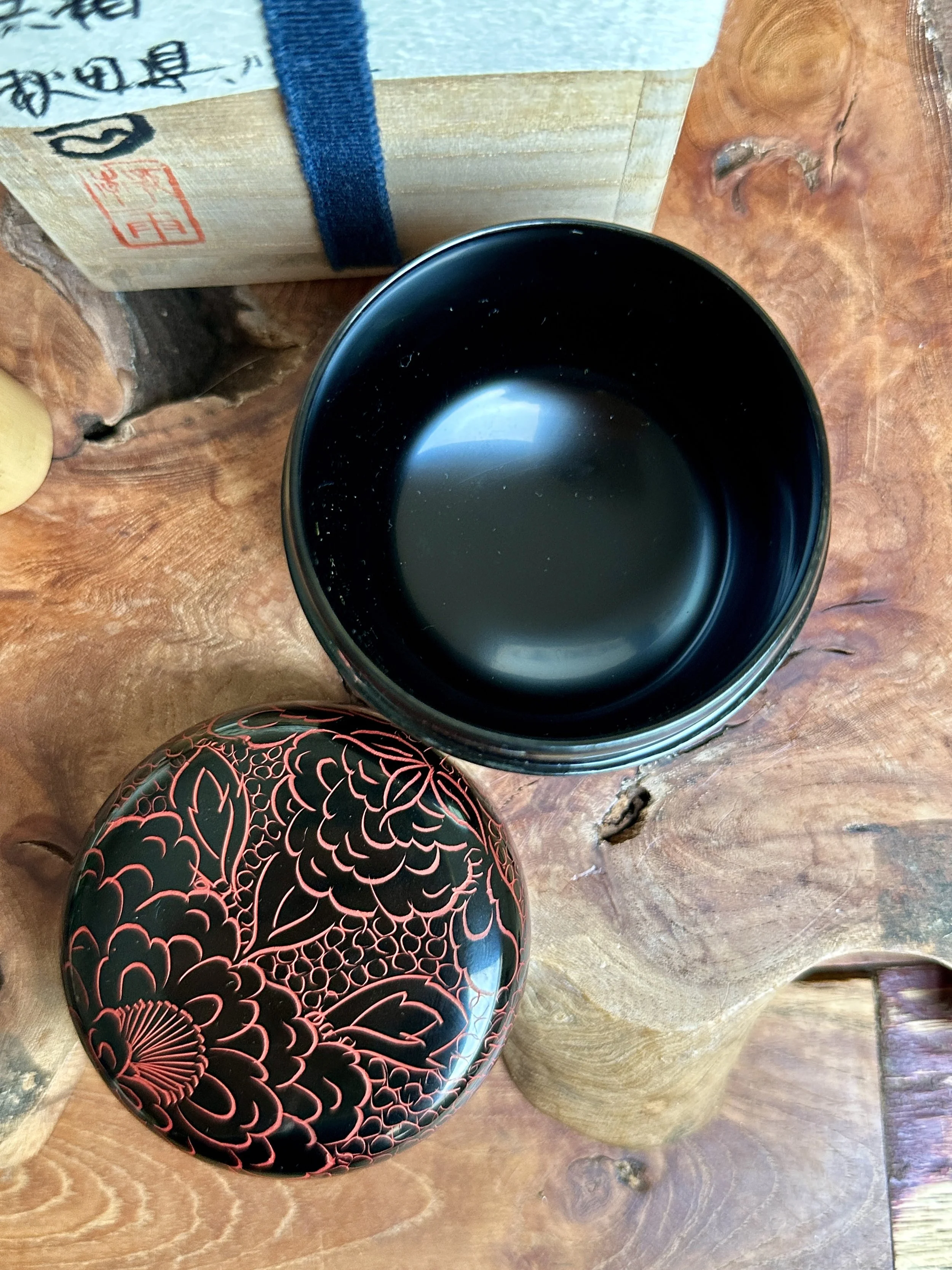 Black lacquered tea caddy, lid off, showing its dark interior. Its red peony-decorated lid rests on a wooden burl. Tomobako box with calligraphy behind. Signed Fujiwara Seiu.