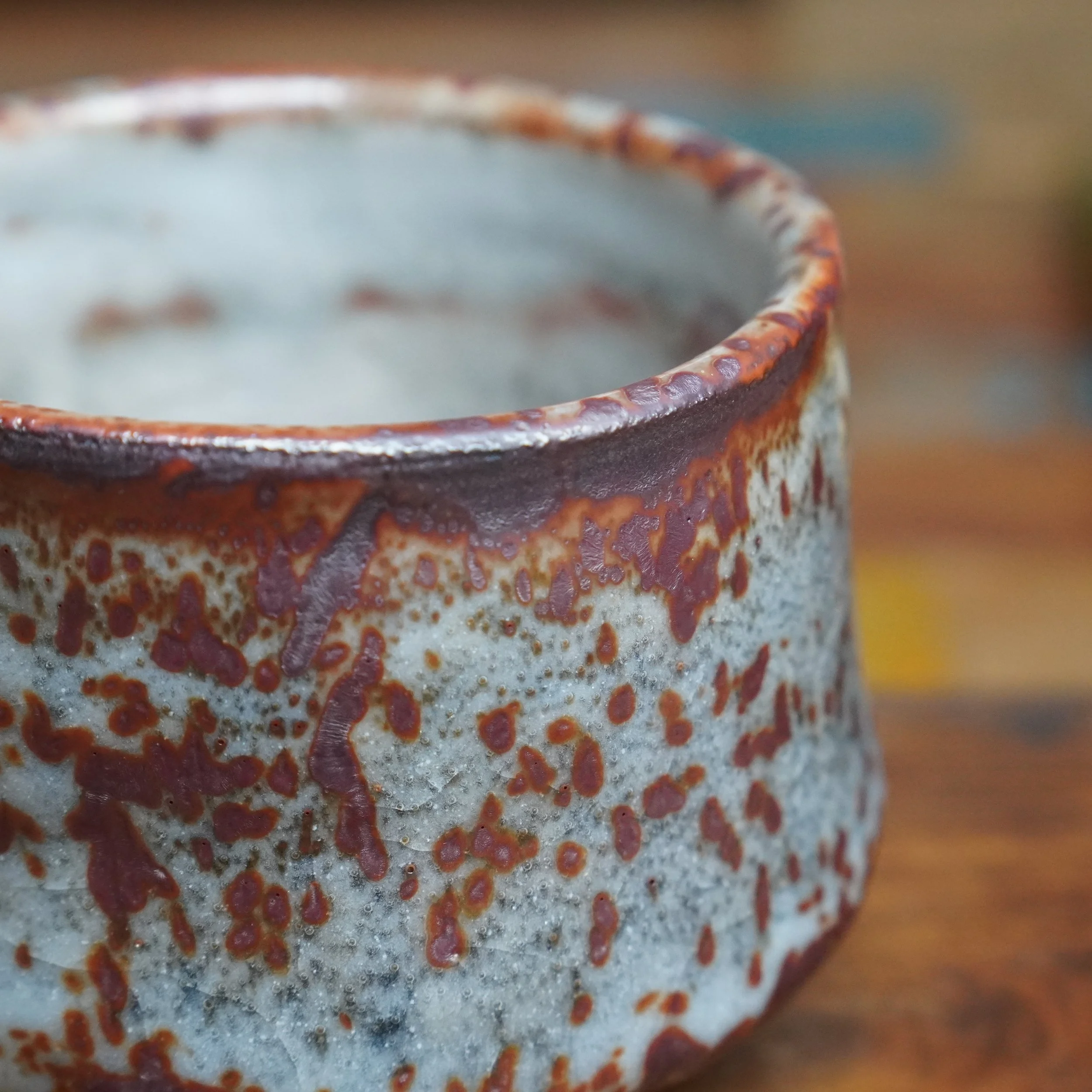 Shino chawan with a coarse pale grey exterior, covered in irregular dark red glaze patches. The top rim features a thick, glossy reddish-brown glaze.