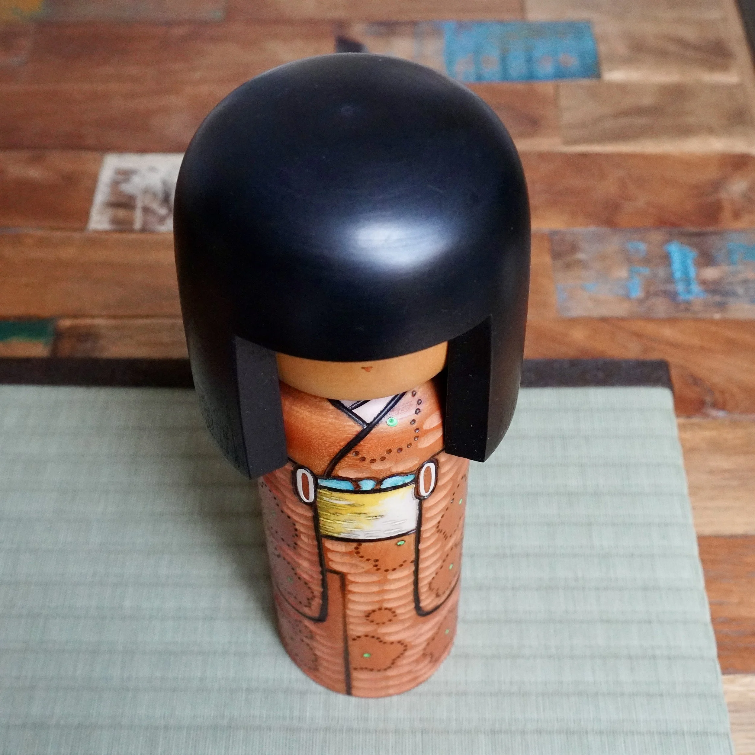 Kisaku kokeshi doll with black hair. Her terracotta kimono features carved textures, painted ume blossoms with green dots, and a pale obi with a turquoise band.