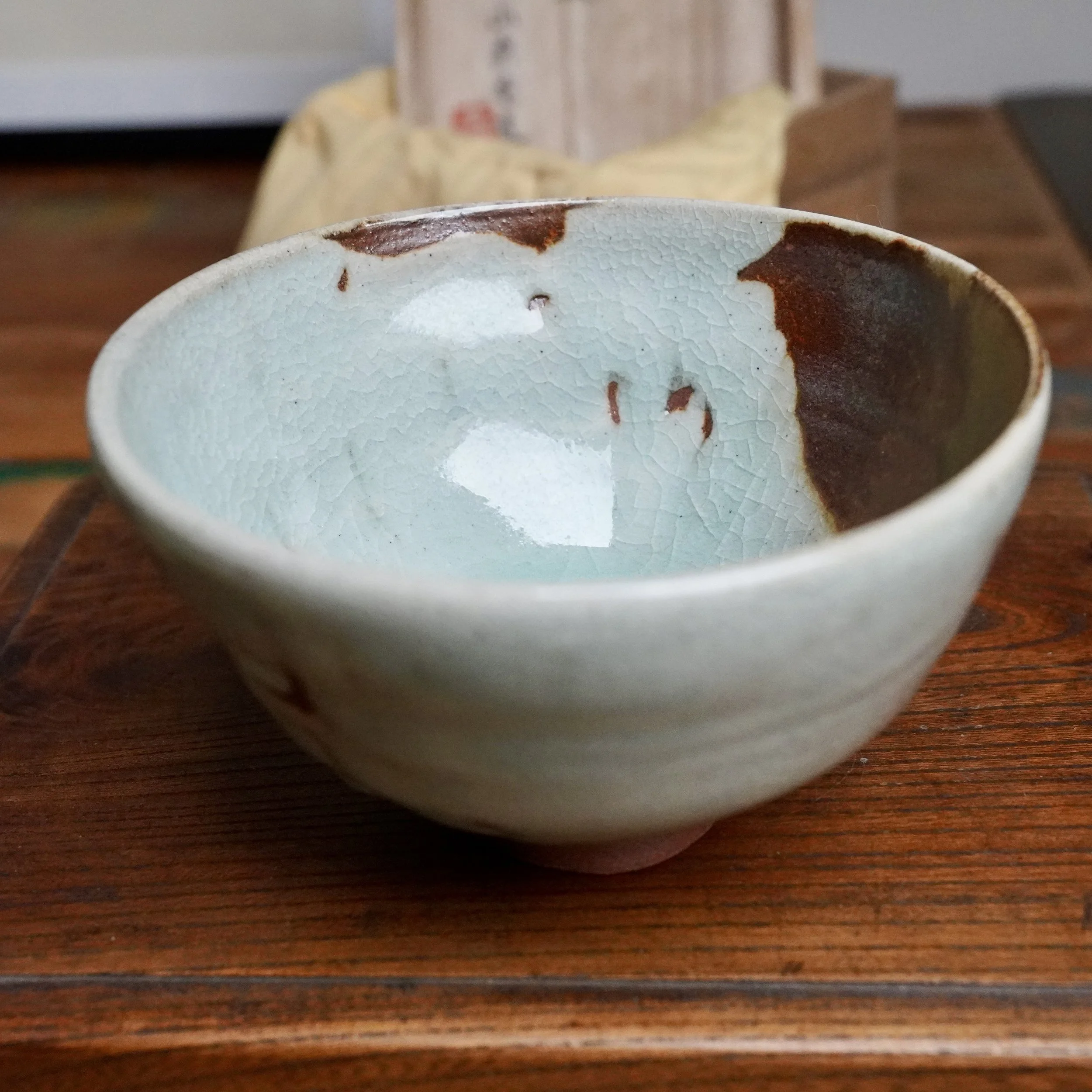 Sansōan chawan with a pale, crackled blue-grey interior glaze and deep rust-brown glaze spilling down one side, resting on a wooden table.
