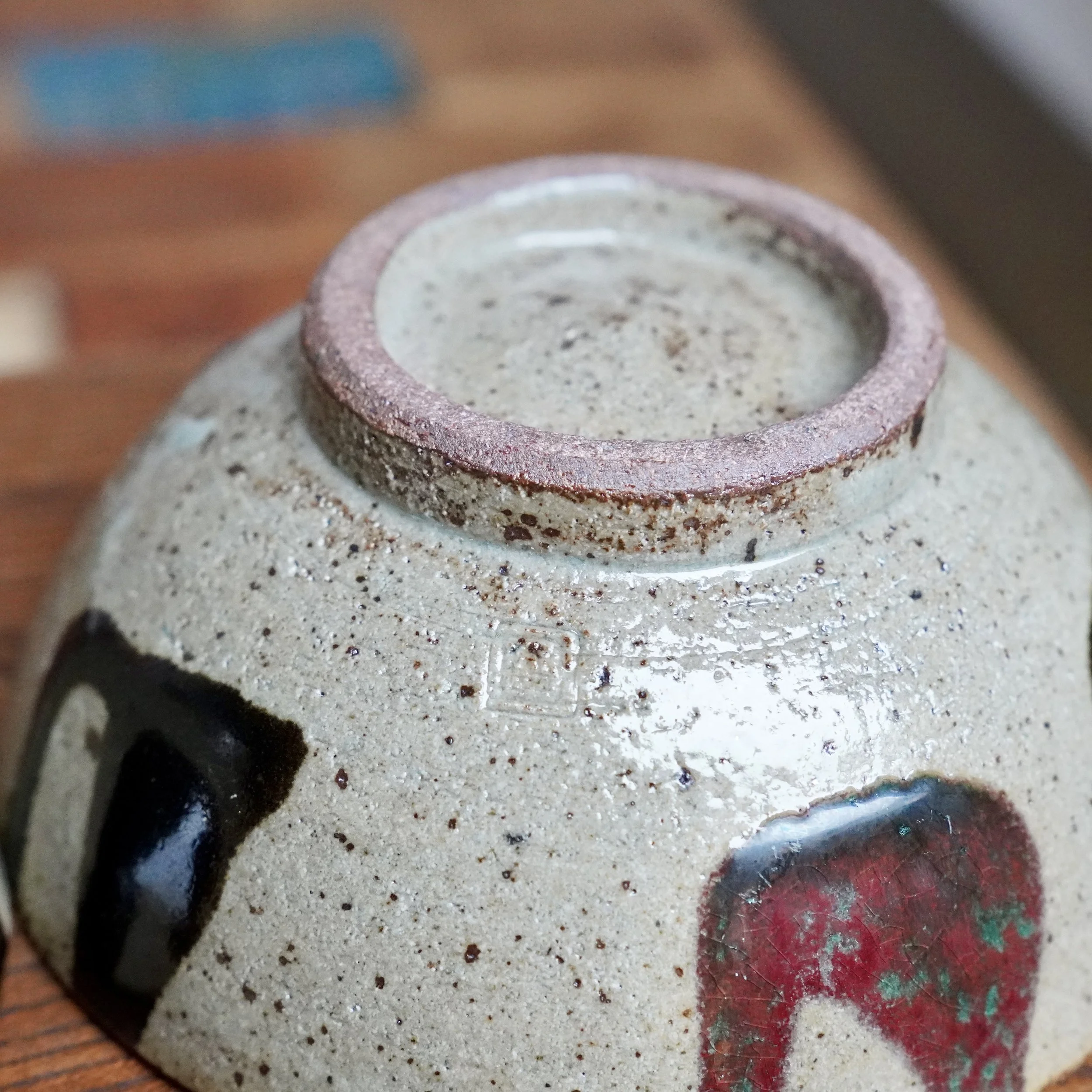 Underside of a Maruta Nobumasa hira-chawan, showing its pale grey speckled clay base, a dark foot ring, and a square maker's mark. Patches of black and crazed wine red glaze are visible on the body.