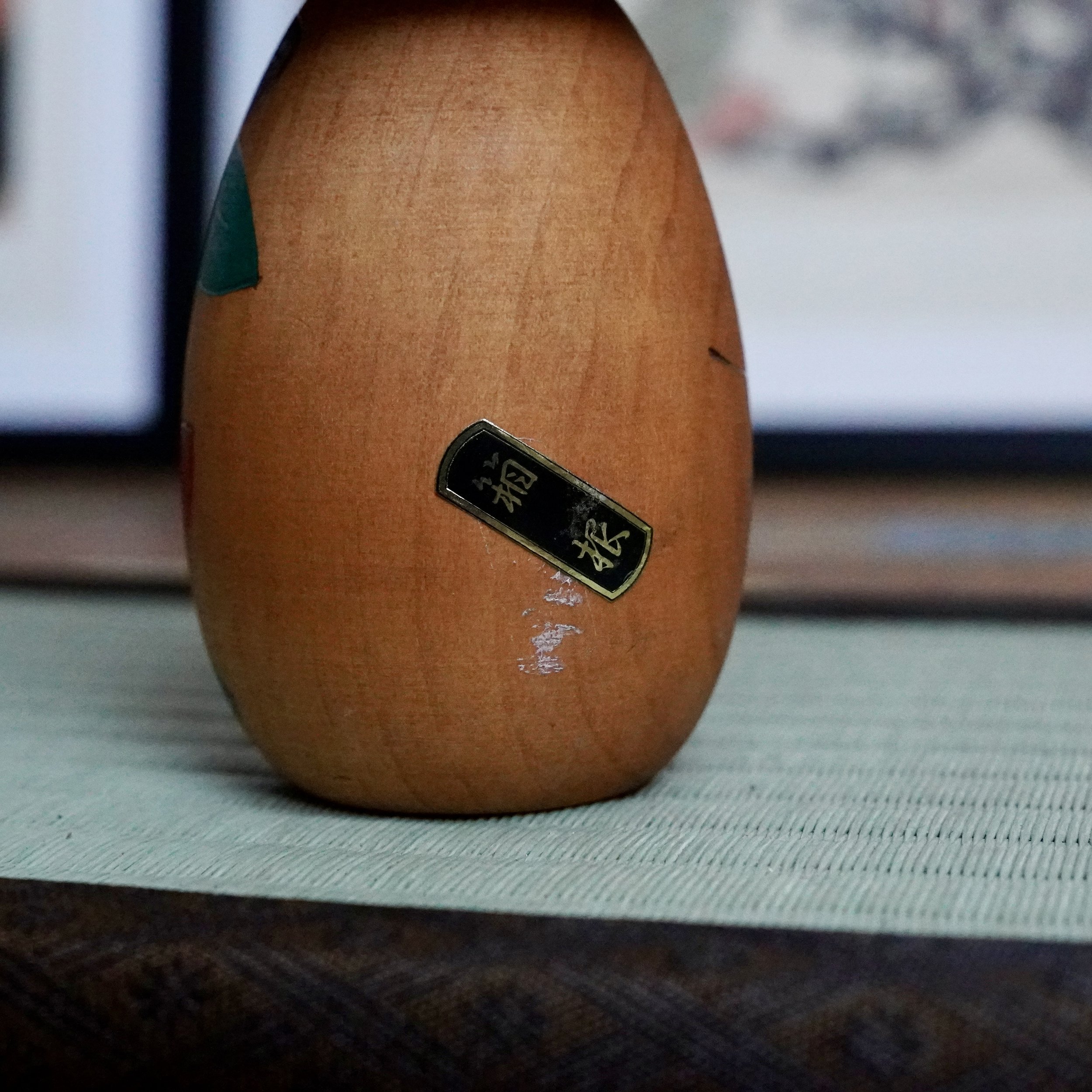 Yuji Kawase kokeshi doll, light brown wooden body, side view. A black label with white Japanese characters and white residue is on its curved side. Doll rests on a woven green mat.
