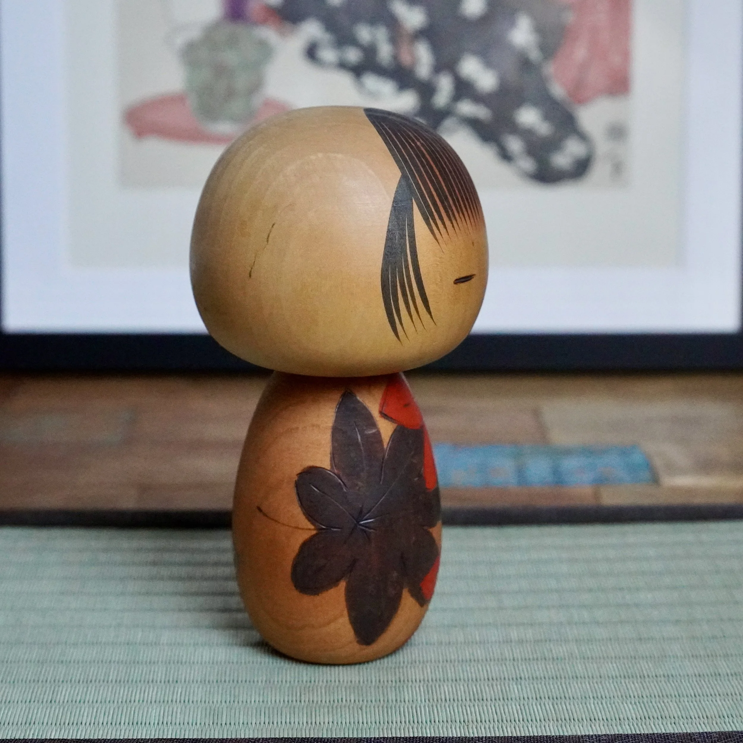 Yuji Kawase sōsaku kokeshi, side view. Light wood doll with carved dark maple leaves highlighted with red, black painted eye, and hair. Blurred art print and tatami mat background.