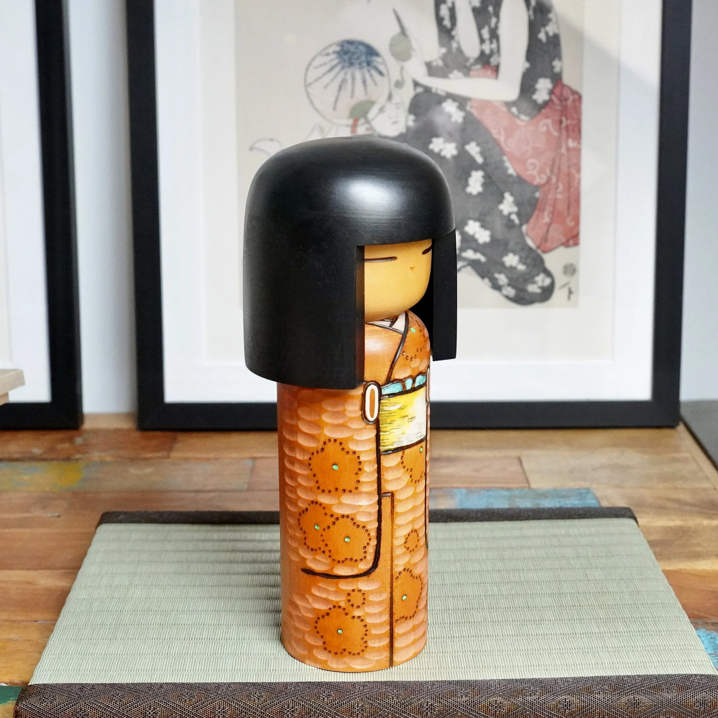 Kisaku sōsaku kokeshi, side view. Black hair, pale face, orange kimono with carved texture, painted ume blossoms, yellow and turquoise obi, on a woven mat.