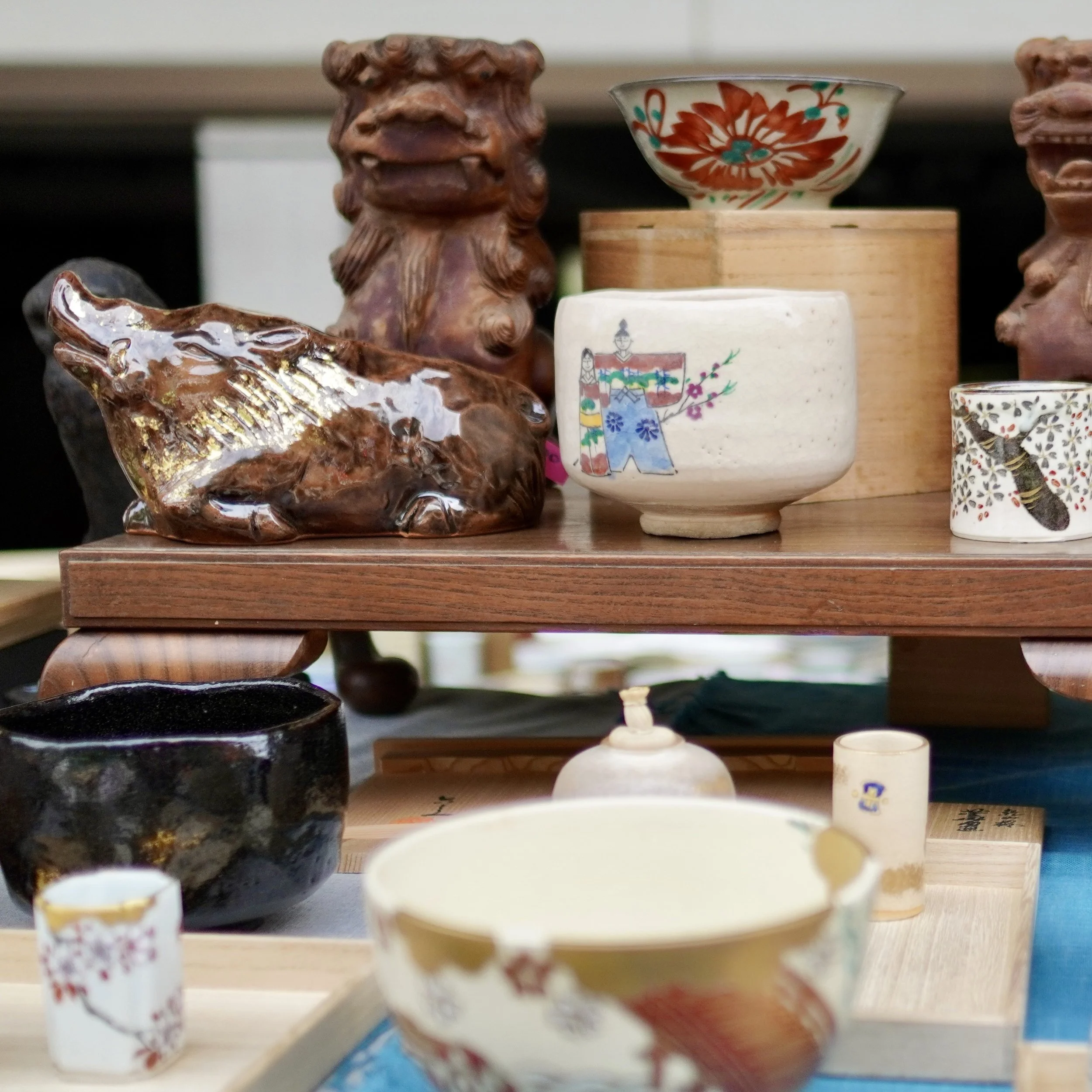 A close-up of a white chawan matcha bowl featuring a hand-painted illustration of figures in traditional dress, placed on a wooden riser next to a brown glazed ceramic animal figure with gold-toned details.