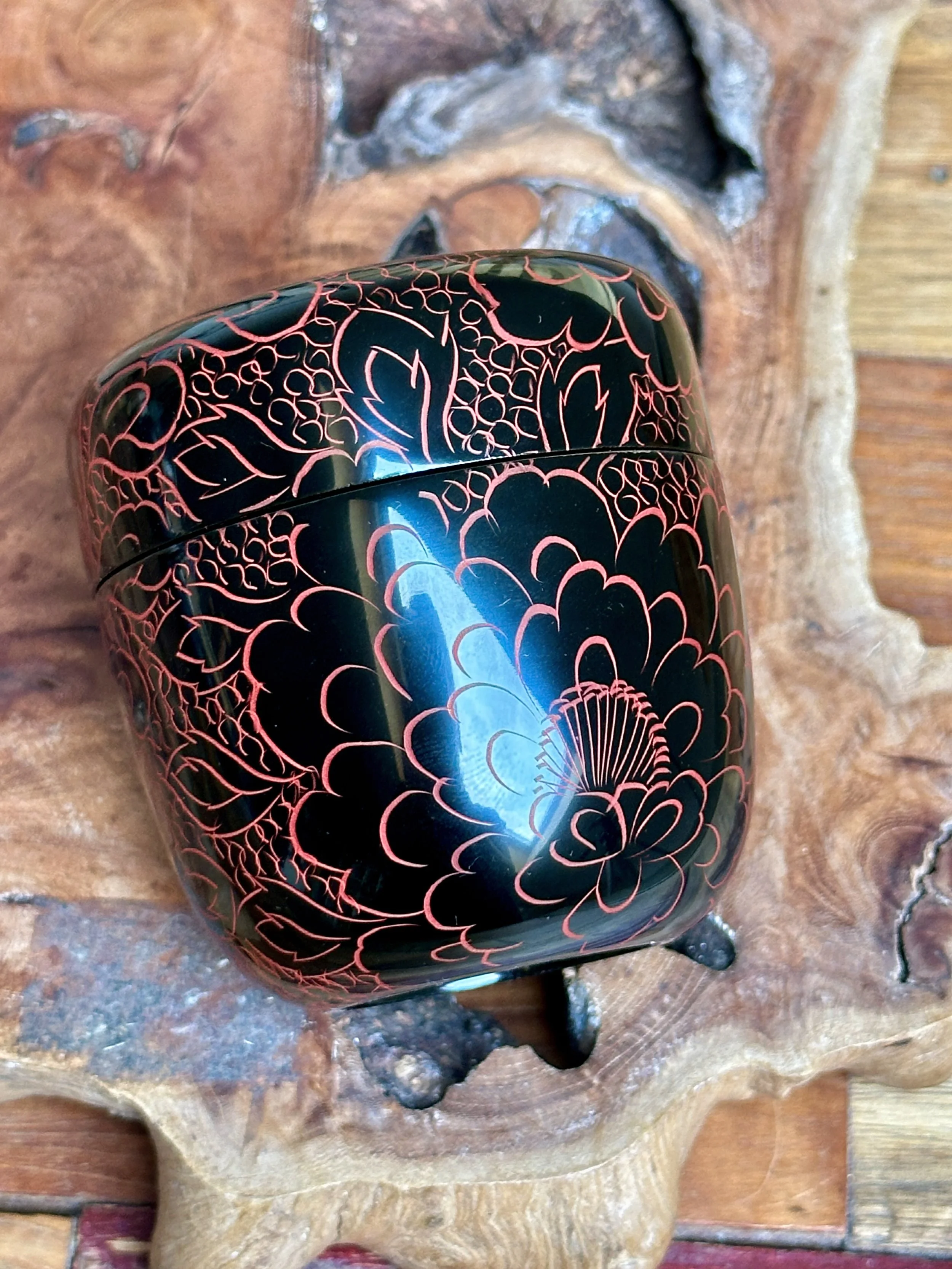 Glossy black lacquered tea caddy with vermilion engraved floral patterns across its rounded body and lid, seen on a rustic wooden slab.