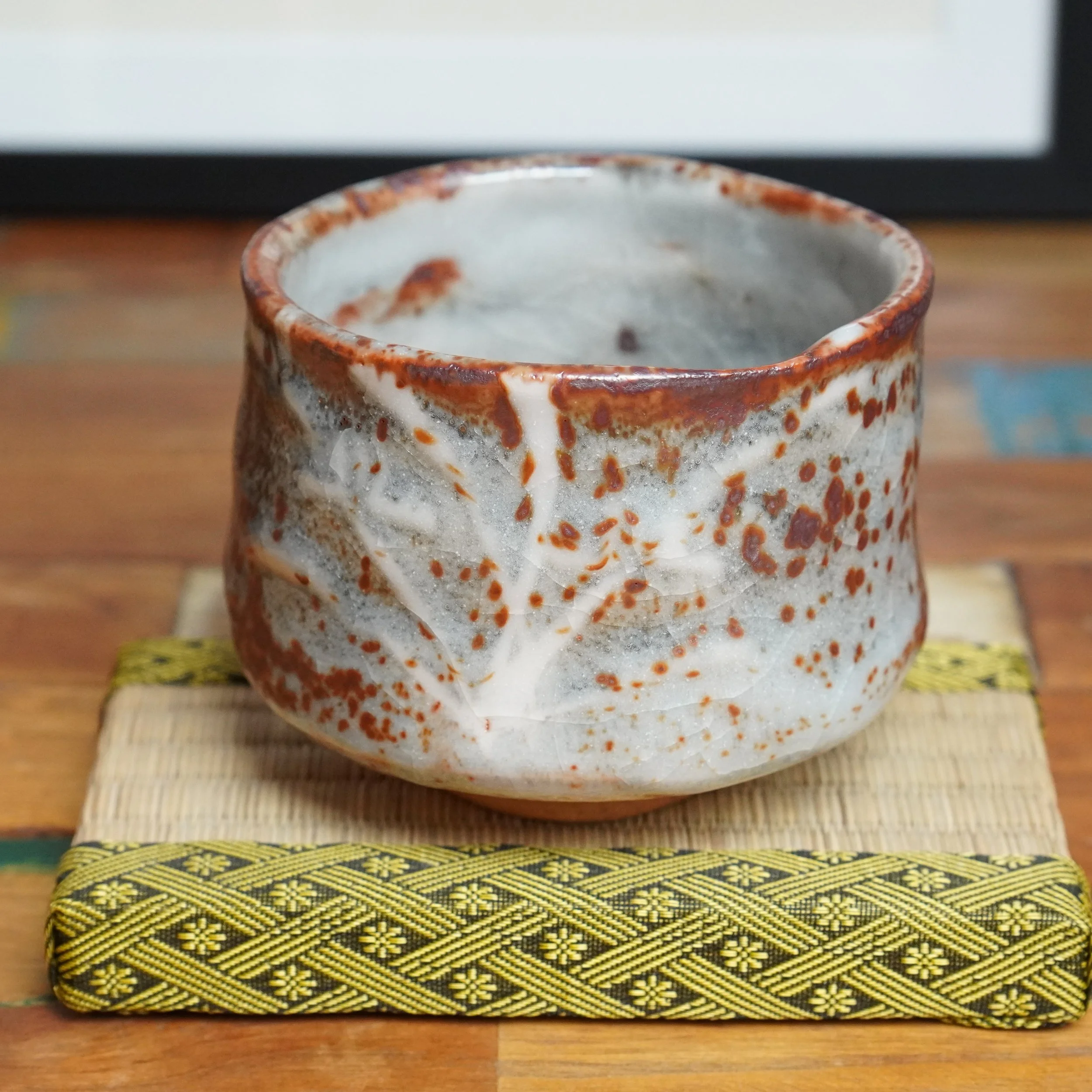 Thick-walled chawan with white and pale grey glaze, featuring deep red speckles and a pale branch-like line, on a woven mat with a yellow and black geometric border.