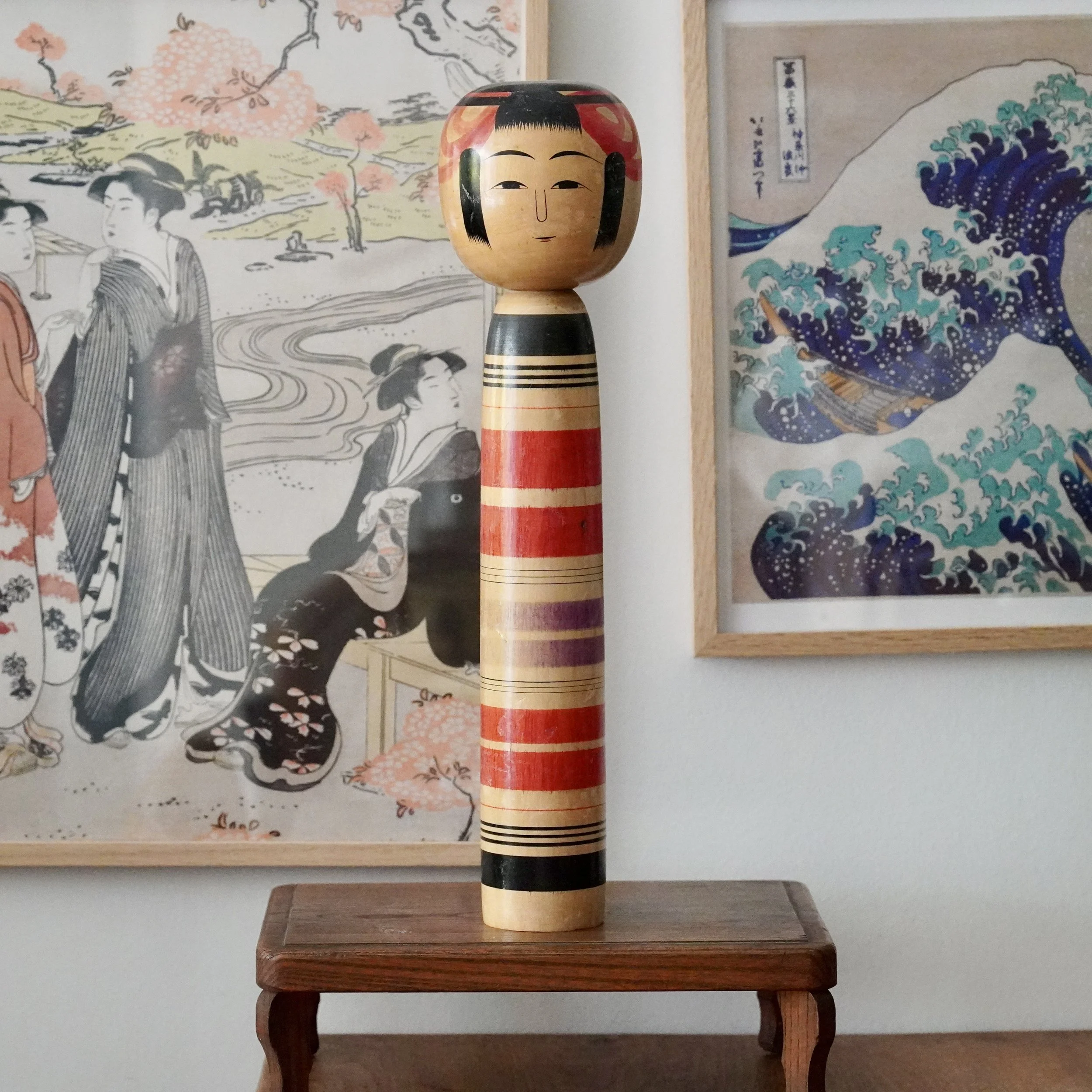 Tsuchiyu kokeshi by Abe Ichirō with red, black and faded purple lathe bands, 36 cm, on wooden stand, woodblock prints behind