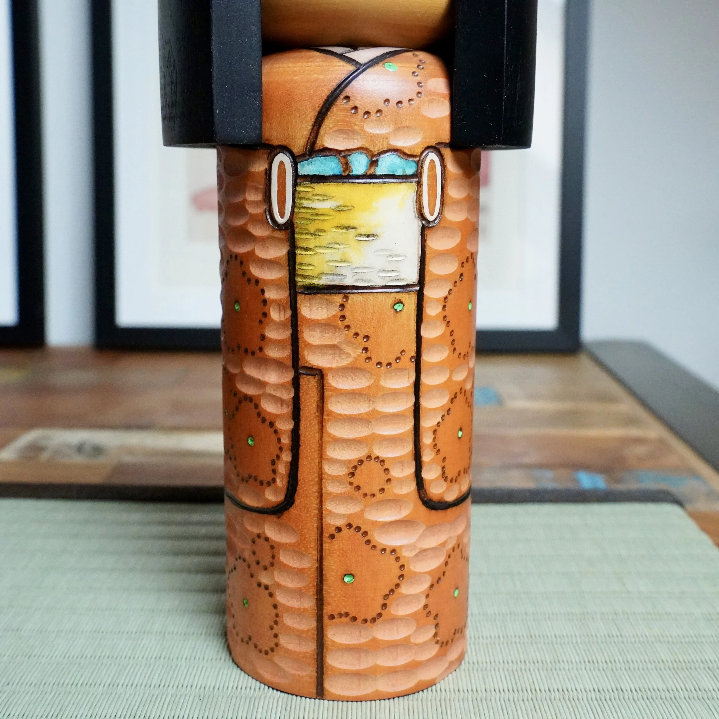 Kisaku sōsaku kokeshi, front view of its orange-brown carved kimono with textured patterns, green dots, and a yellow-white obi with turquoise accents.