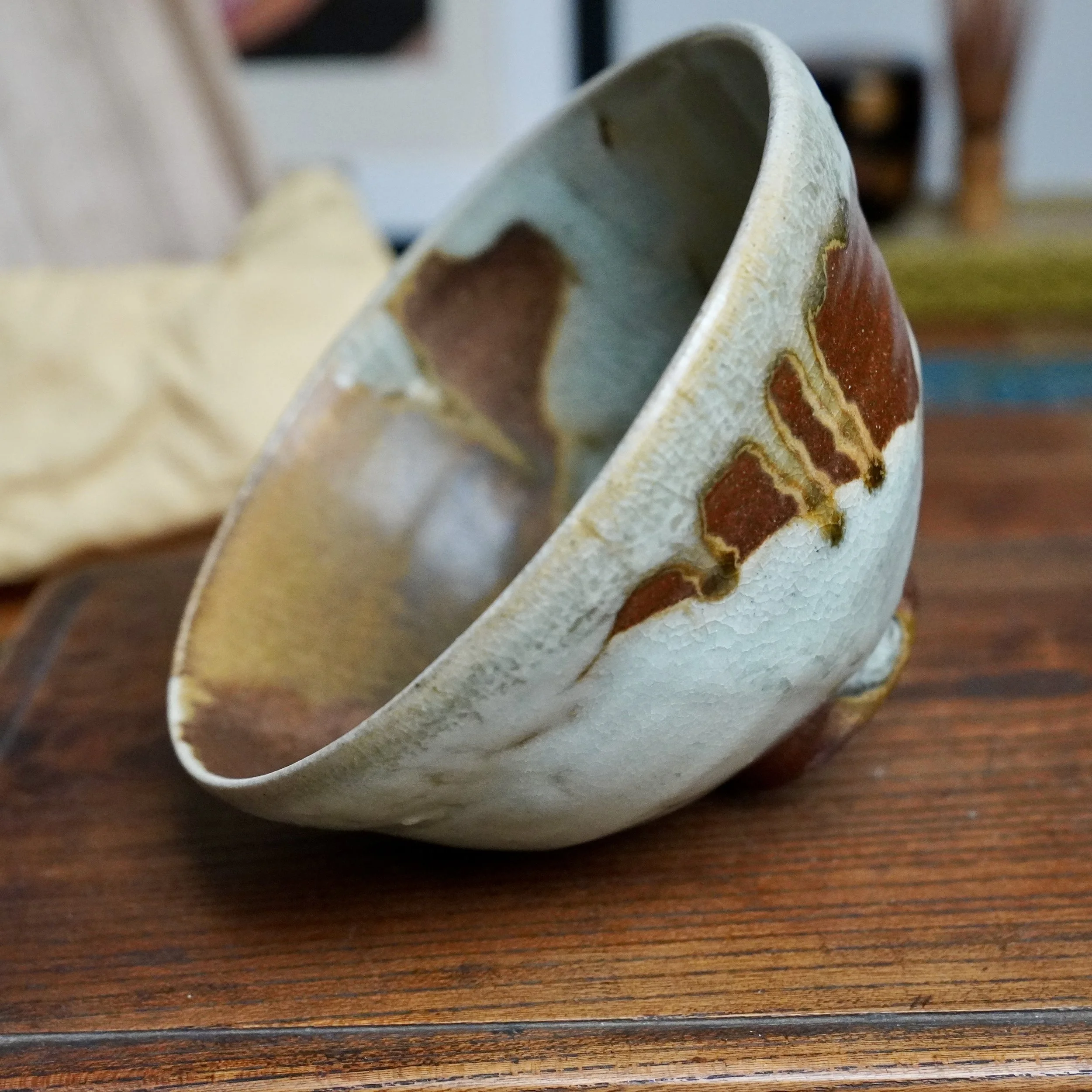 Ceramic chawan tilted on a wooden surface, displaying pale blue-grey crackled glaze with rust-brown and ochre glaze flowing down its side.