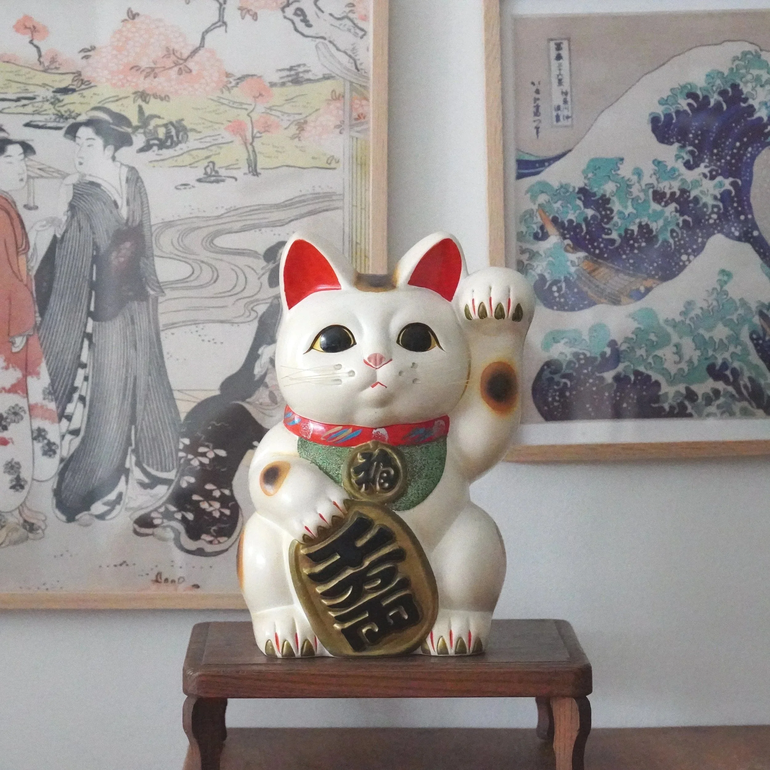 A wooden table with a Japanese maneki neko cat figurine with red ears and black eyes, and a black and gold coin-shaped object with Japanese characters. There are traditional Japanese ukiyoe pictures on the wall behind the table.