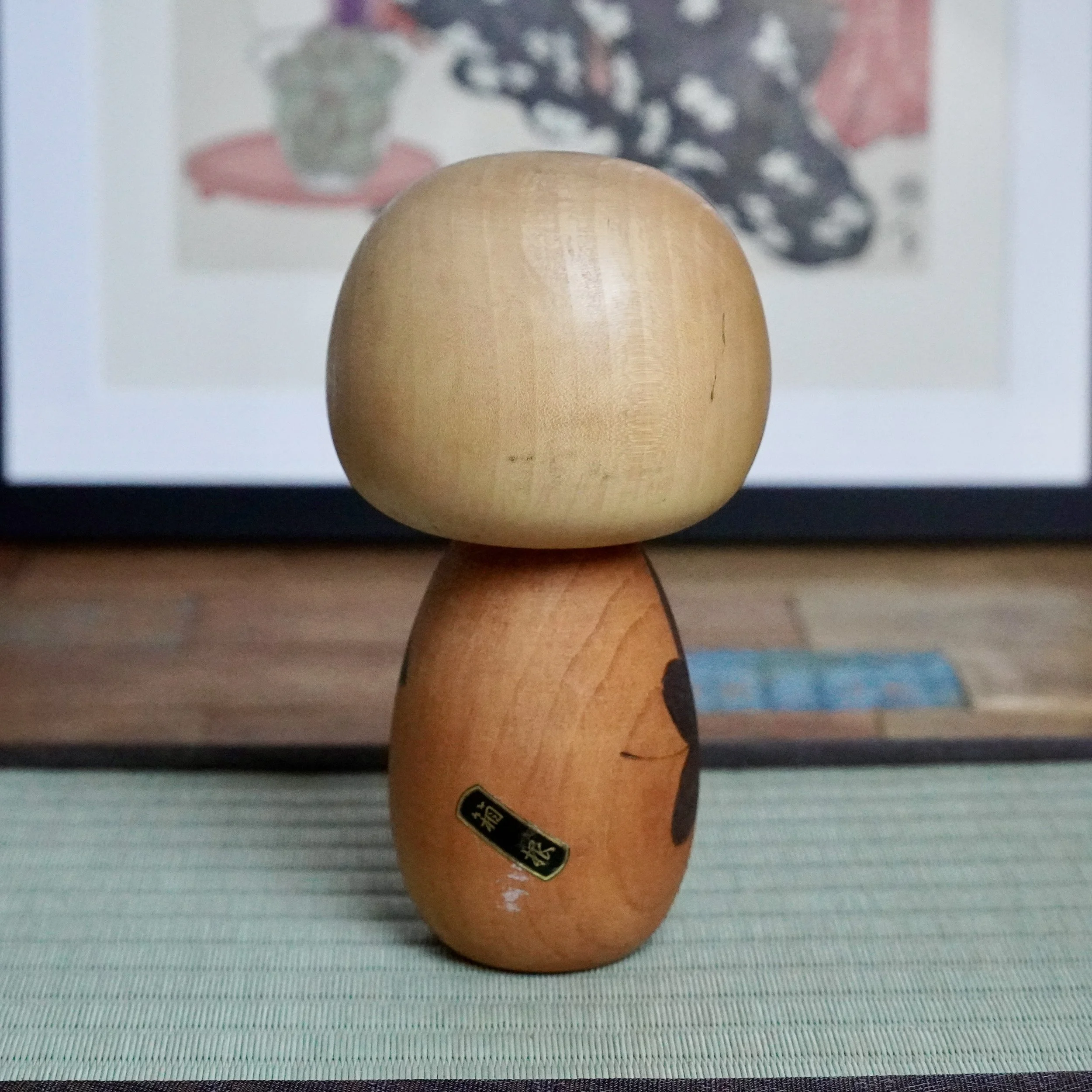 Front view of a Yuji Kawase kokeshi doll, light brown head, medium body carved with dark momiji leaves and a label, on a green woven mat.