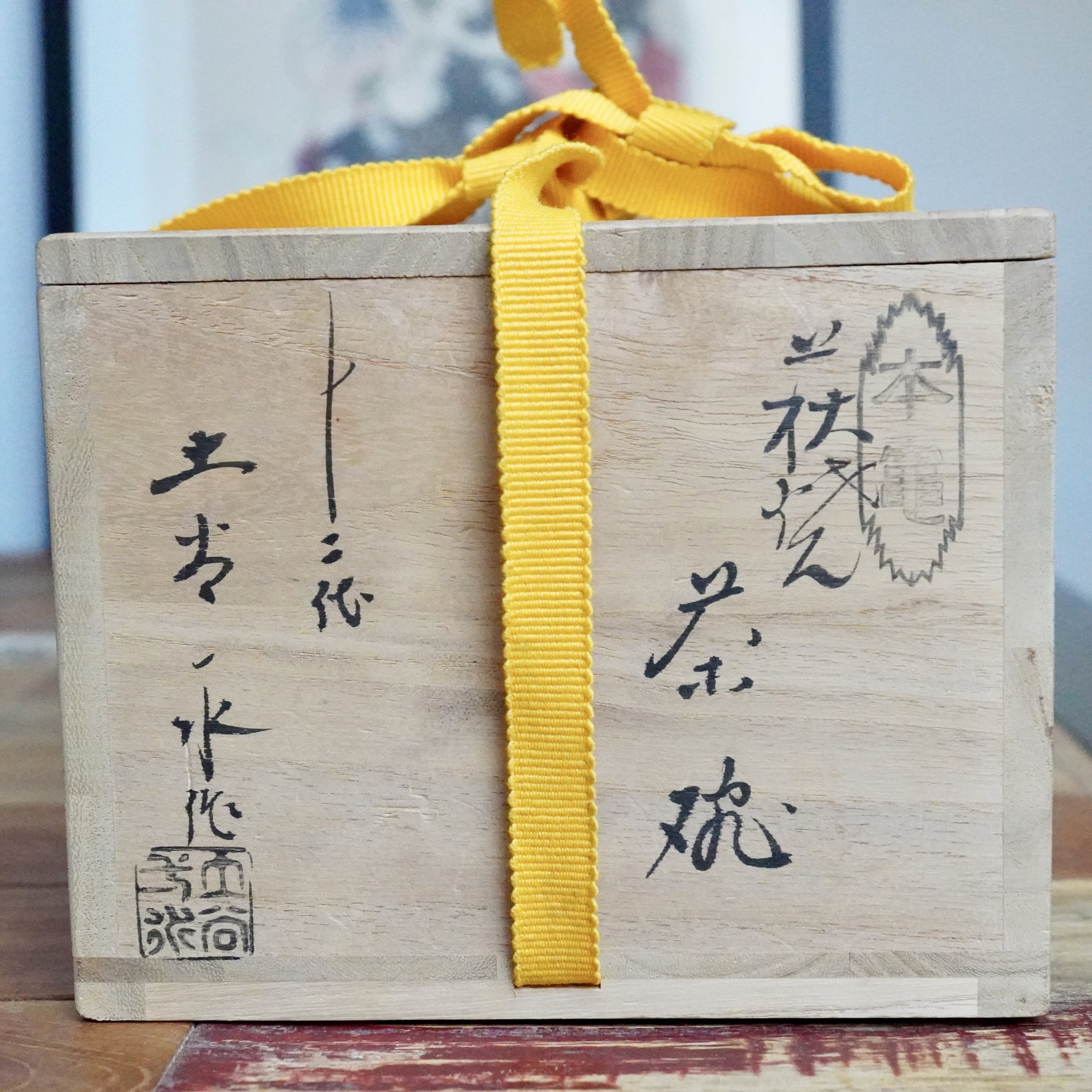 Light wooden tomobako tied with a yellow ribbed ribbon, displaying black Japanese calligraphy, Tsuchiya Issui's name, and a square seal impression.