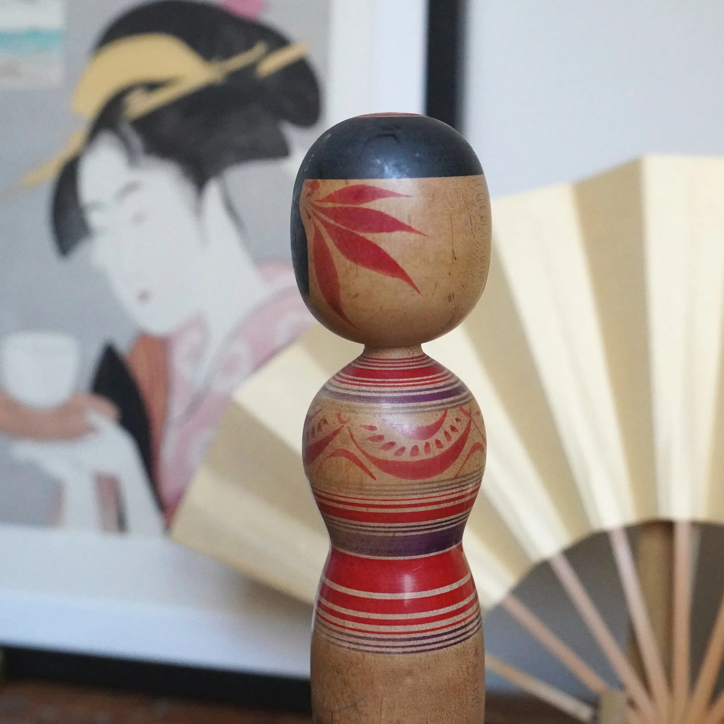 Tsugaru kokeshi by Sasamori Junichi with a black head, red peony motifs, and an hourglass body with red and purple stripes. Blurred painting and fan in background.