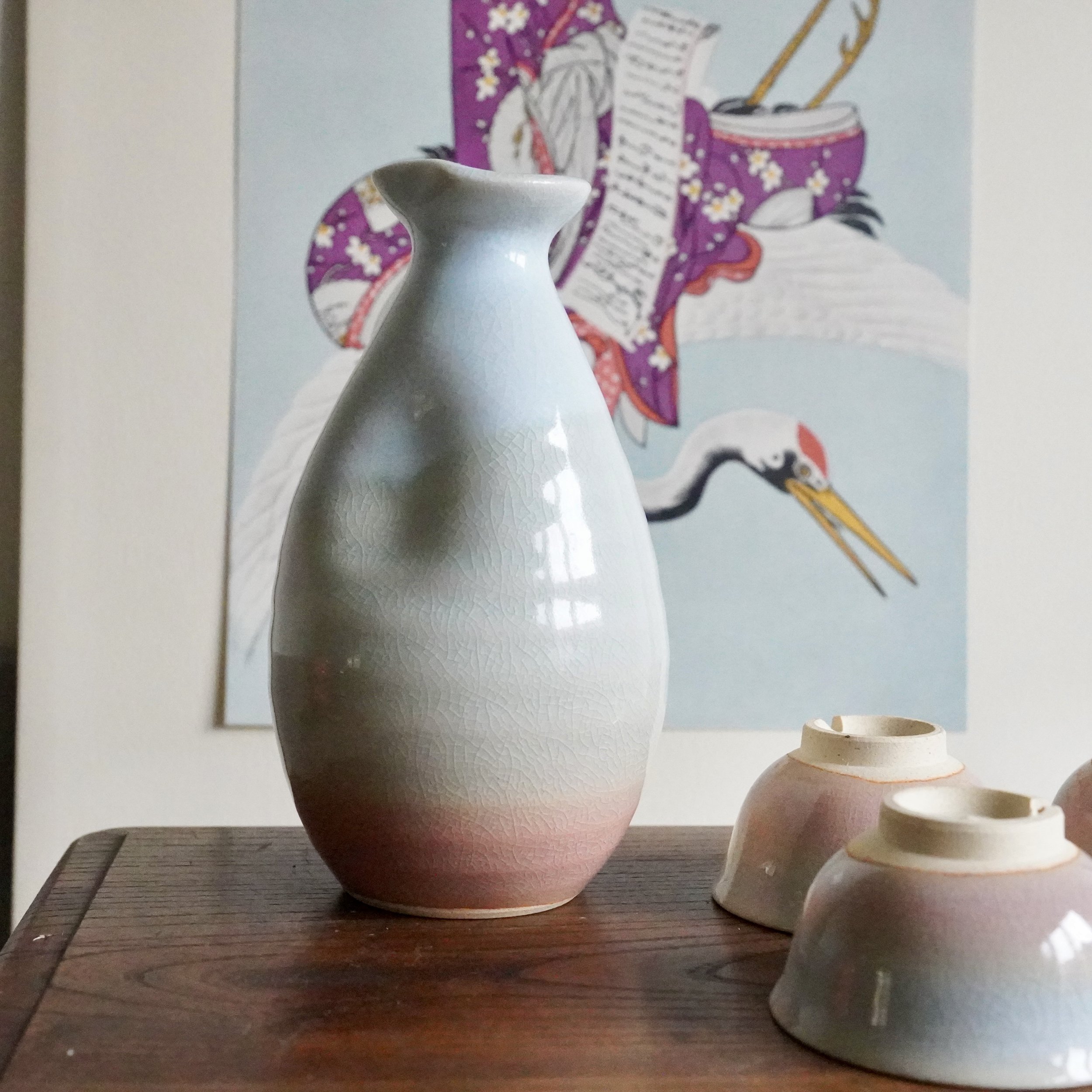 Hagi-yaki sake tokkuri and several ochoko, all with a crackle glaze blending from soft pink to pale blue-grey, on a dark wood surface before a blurred Japanese crane painting.