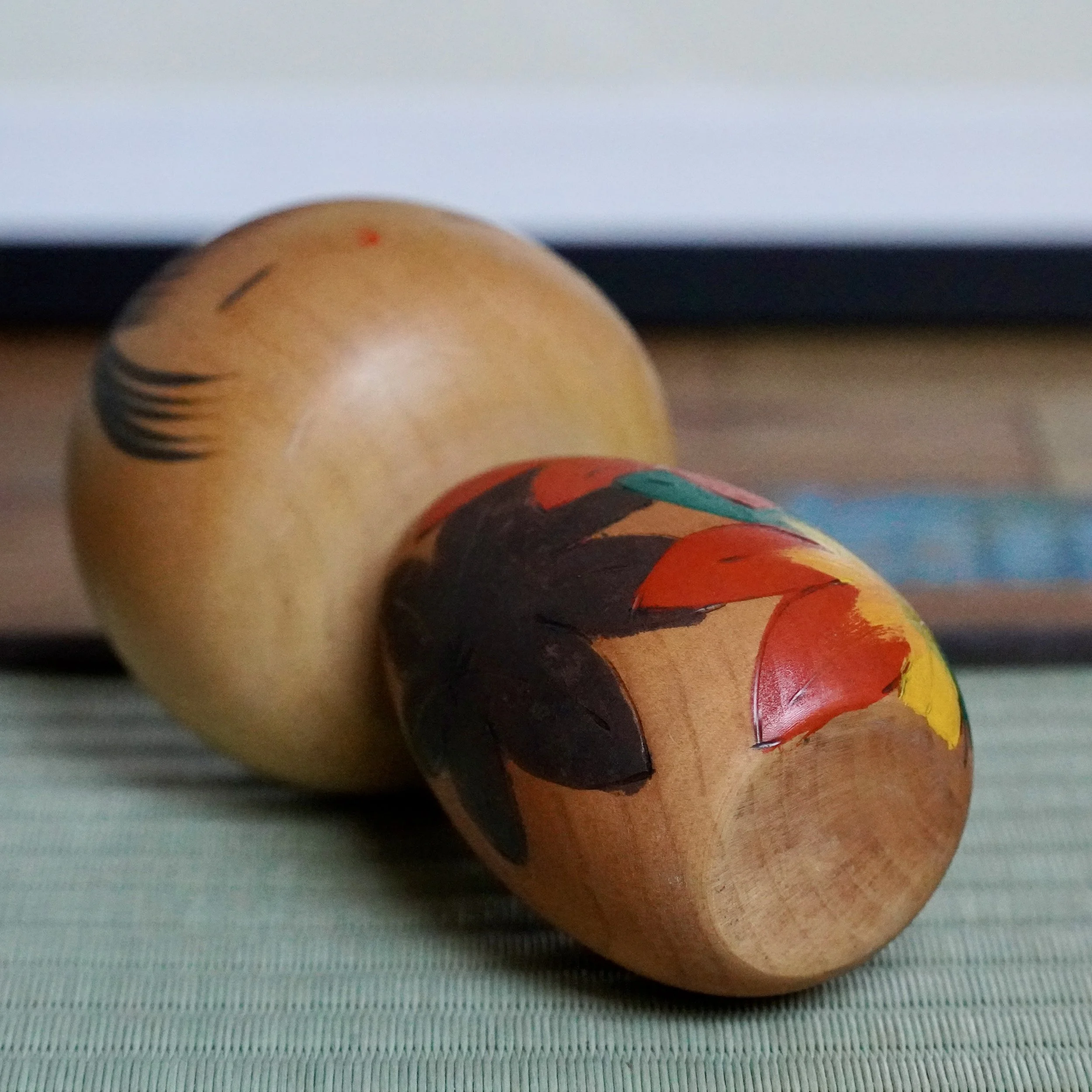 Two Sōsaku kokeshi by Yuji Kawase on a mat. One large doll has black markings, the smaller features carved red, dark brown, green, and yellow autumn leaves.