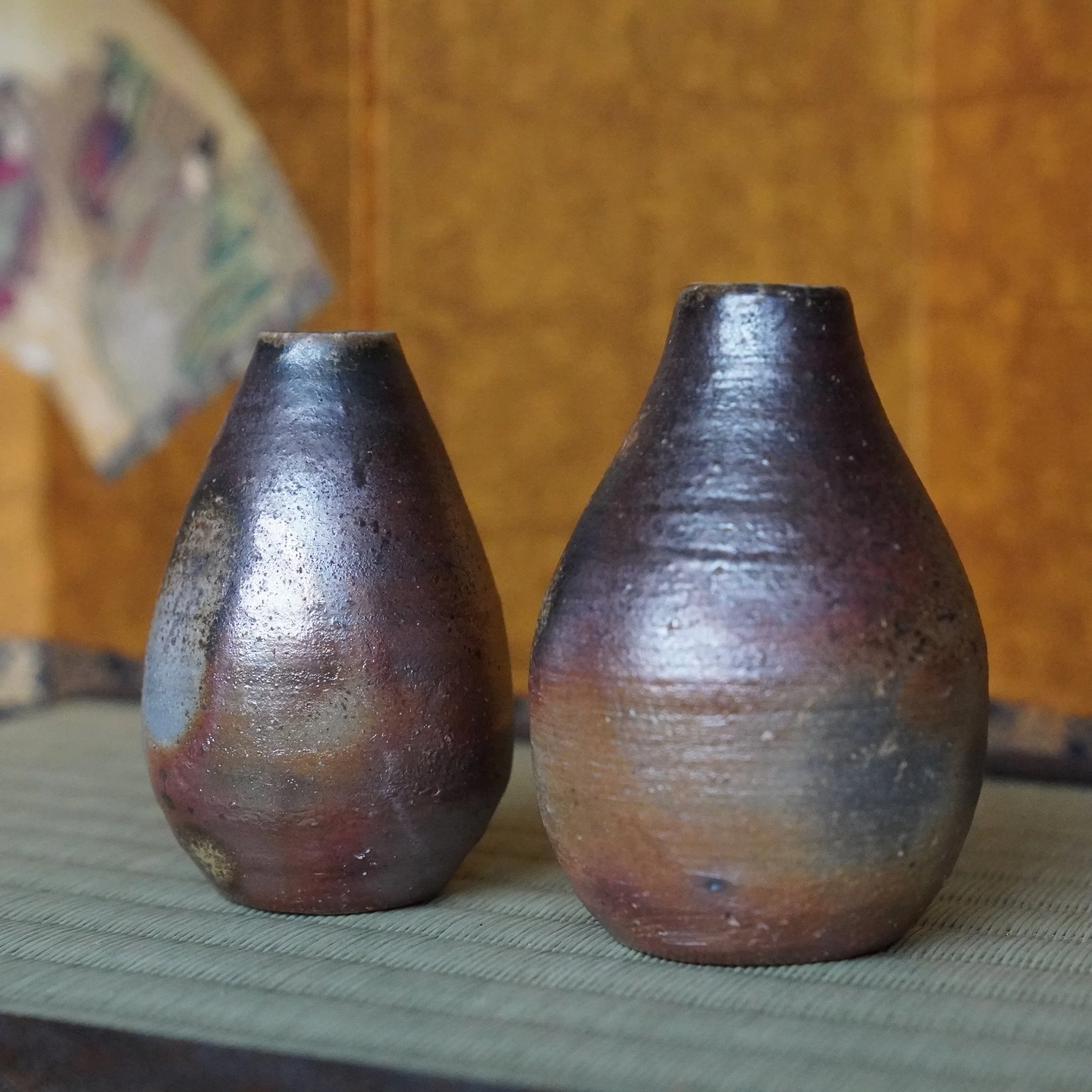 Shibaoka Tōsendō, Kōzan-gama Bizen-yaki tokkuri vases. Left dark, metallic, right reddish-brown, grey base. On a woven mat.