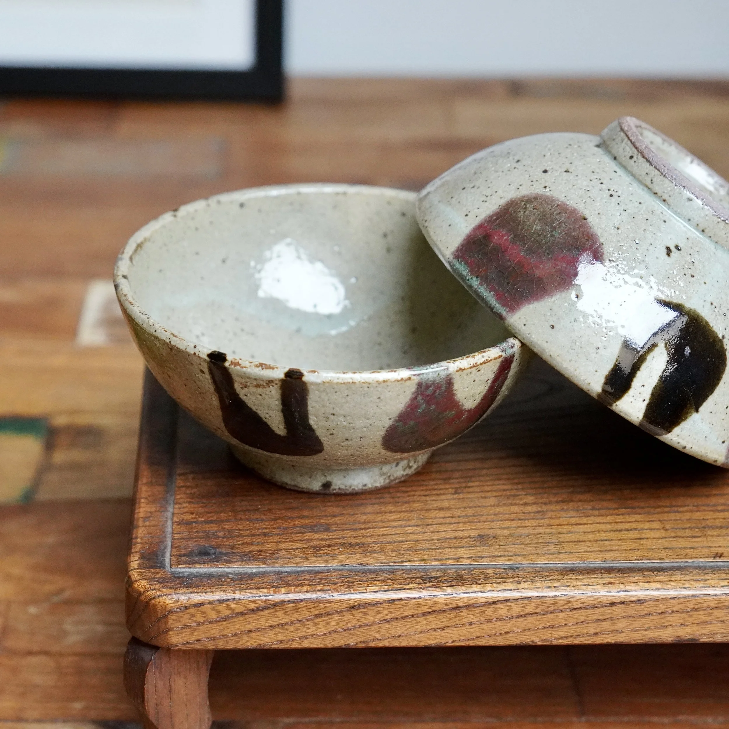 Two Maruta Nobumasa hira-chawan bowls. One upright, the other inverted, showing speckled pale grey clay with milky white, dark brown, and wine red glaze on a wooden tray.