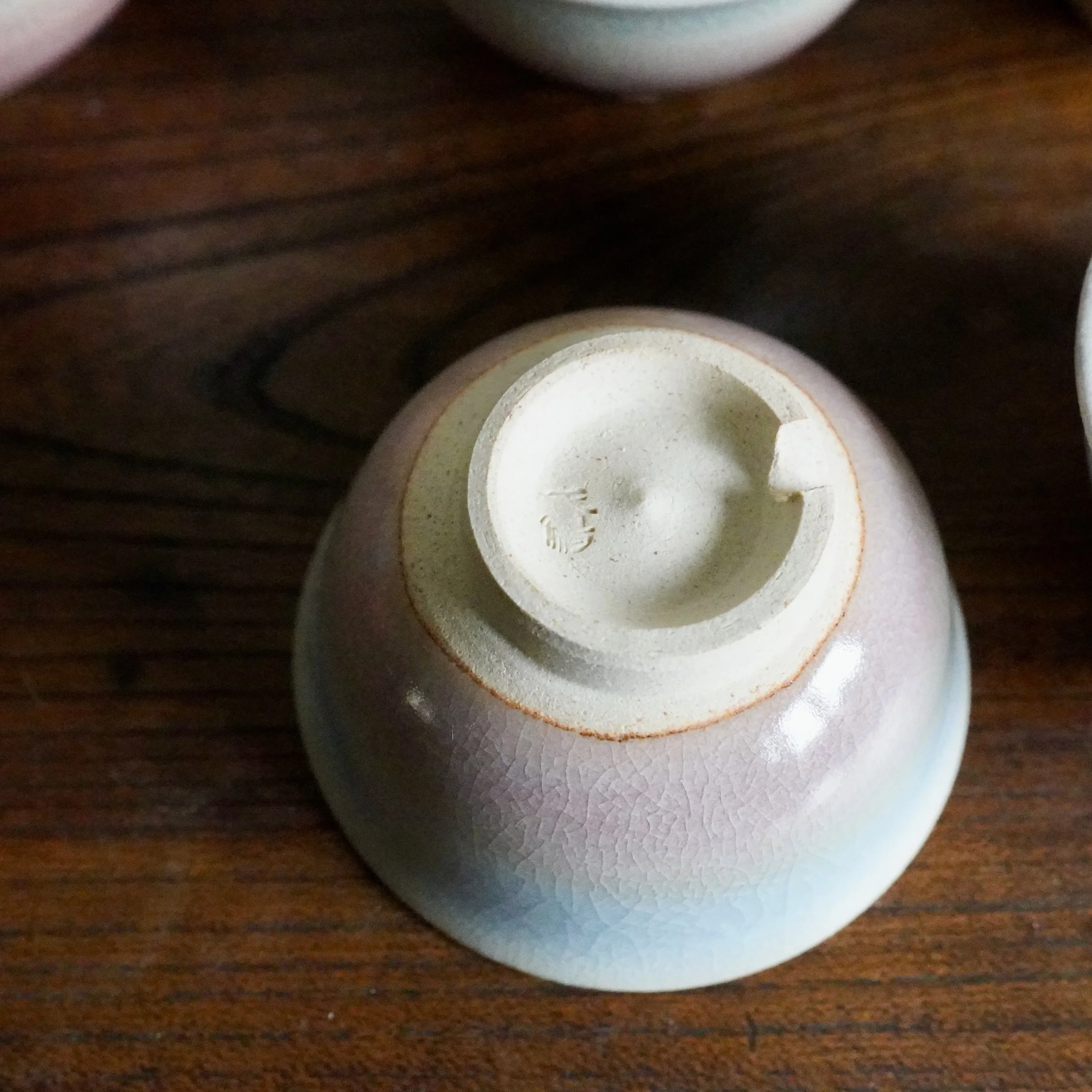 Underside of a Hagi-yaki ochoko. Ceramic cup with a pink-to-blue gradient glaze and fine crazing. Unglazed cream foot shows a kiln stamp. On dark wood.