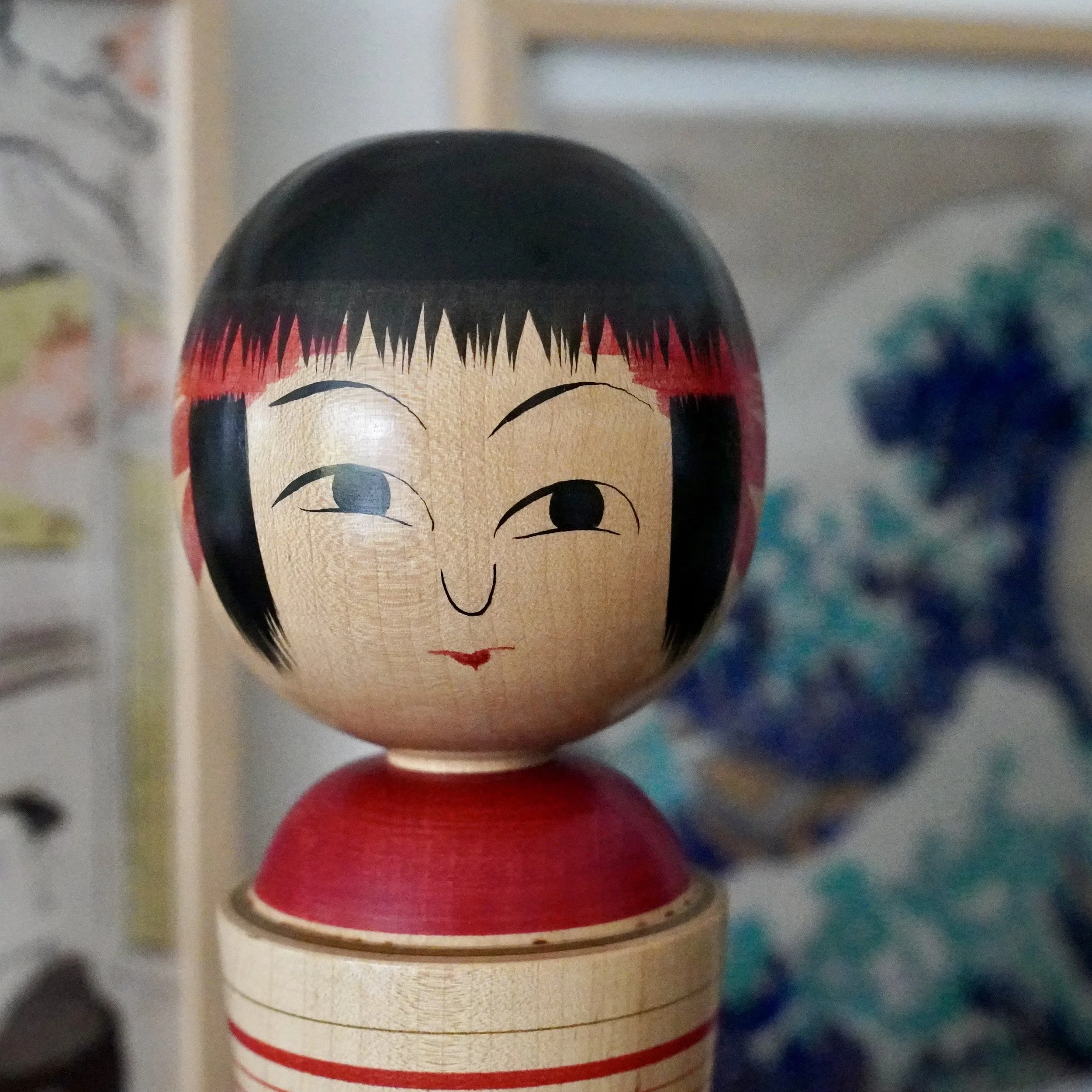 Front close-up of an Izu Mamoru kokeshi doll, featuring a pale wood head with black hair, red fringe, painted eyes, and small red lips. A red collar sits on the light wood body.