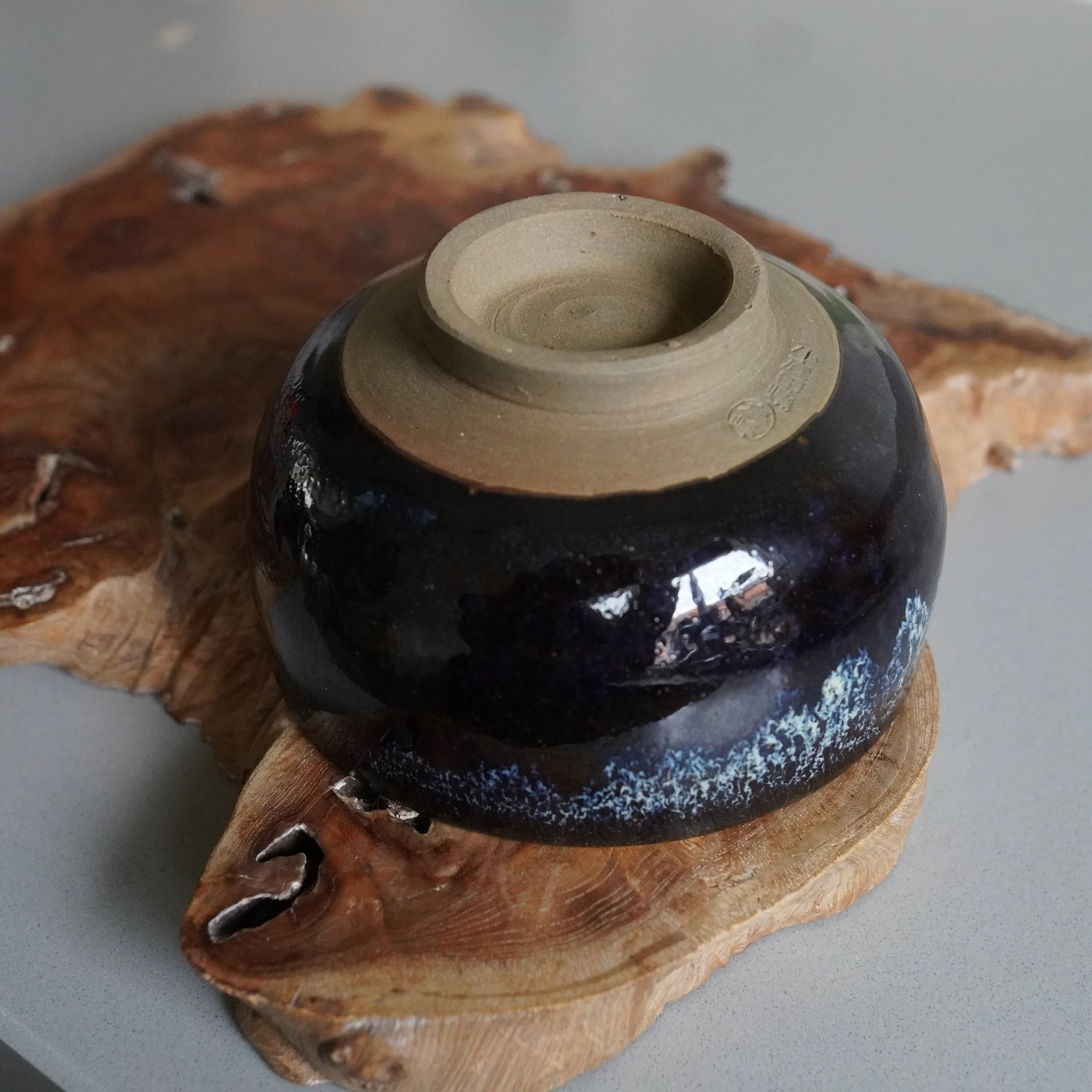 Onta-yaki matcha chawan by Rokuhō, underside view. Dark brown-blue glazed with white speckles, unglazed clay foot with maker's signature, on a wooden slab.