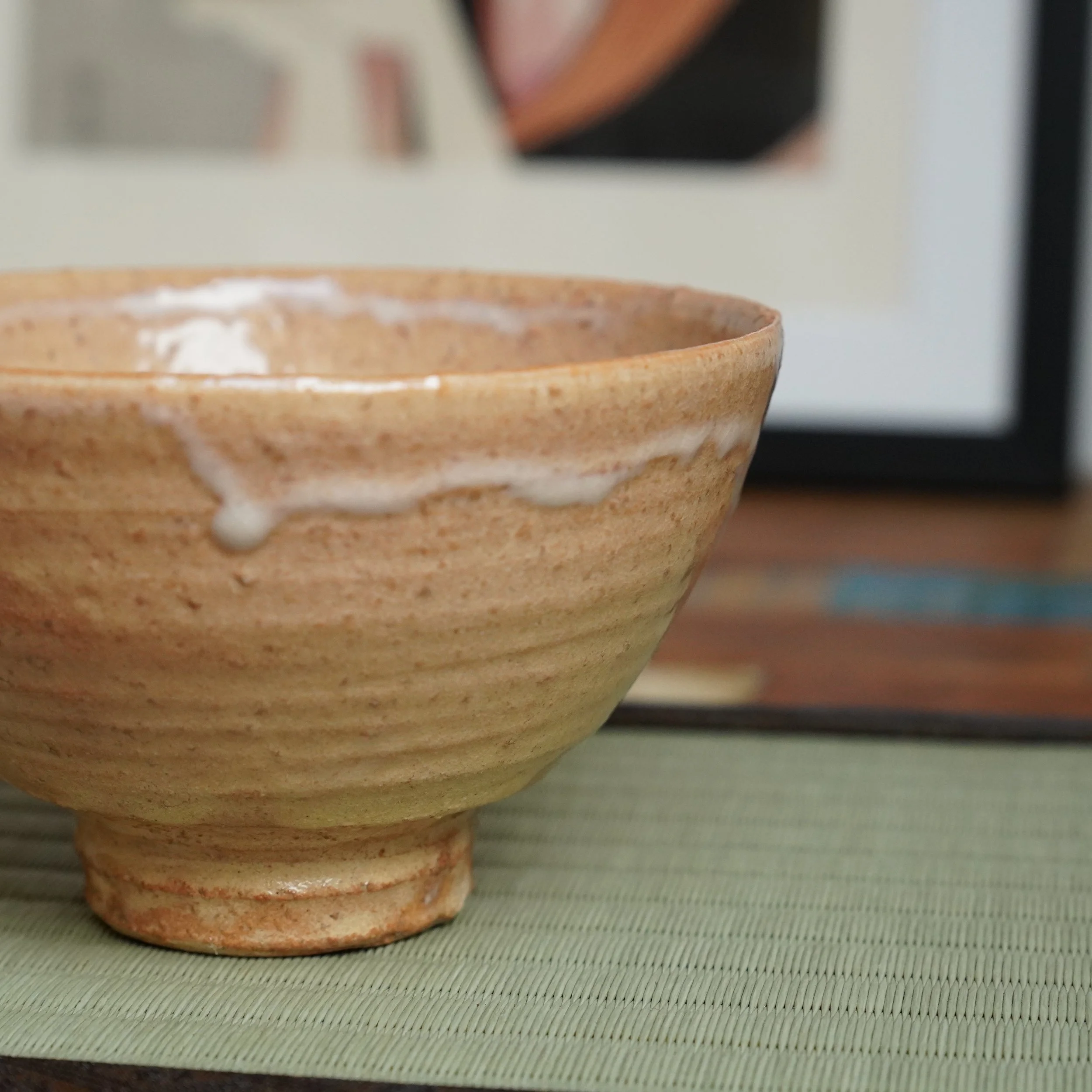 Kaneko Nobuhiko Hagi-yaki chawan, a ribbed, light tan tea bowl with white glaze drips, resting on a green woven mat. Blurred background.