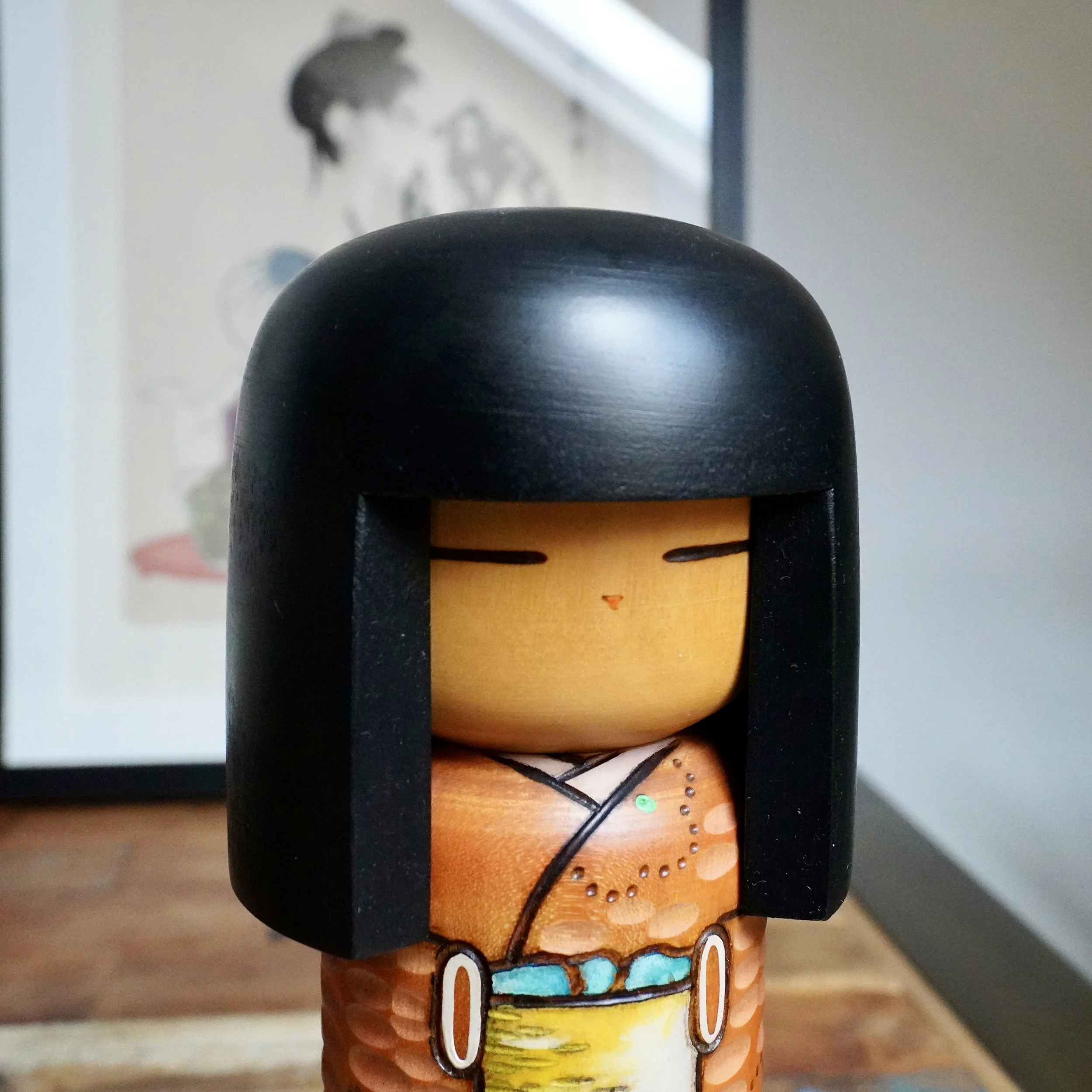 Sōsaku kokeshi by Kisaku with black bob hair, light wood face, and an orange kimono carved with scooped indentations and painted ume blossom outlines.