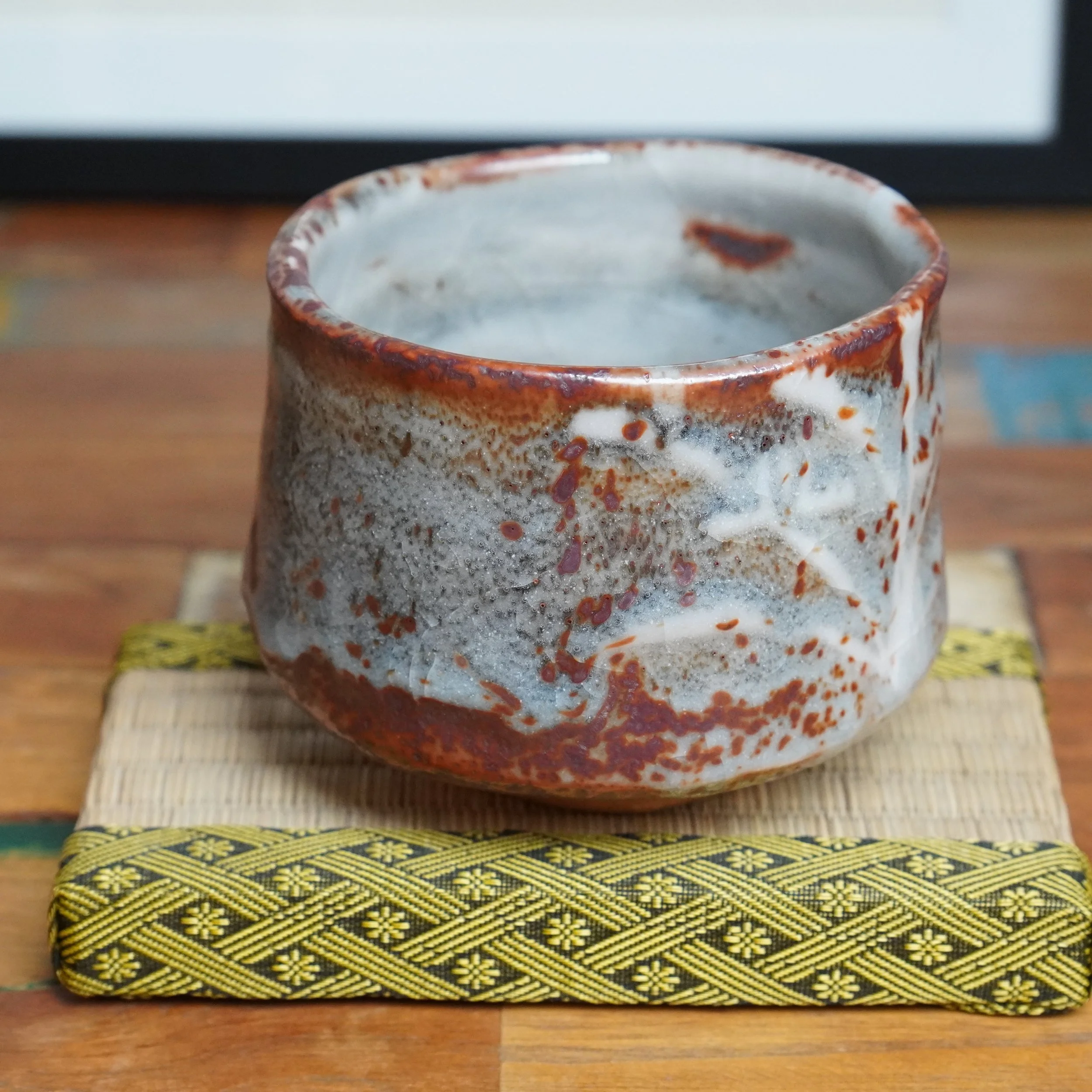 Ceramic chawan with patchy white, grey, and reddish-brown speckled glaze, on a light woven mat with a black and yellow geometric border, sitting on a wooden surface.