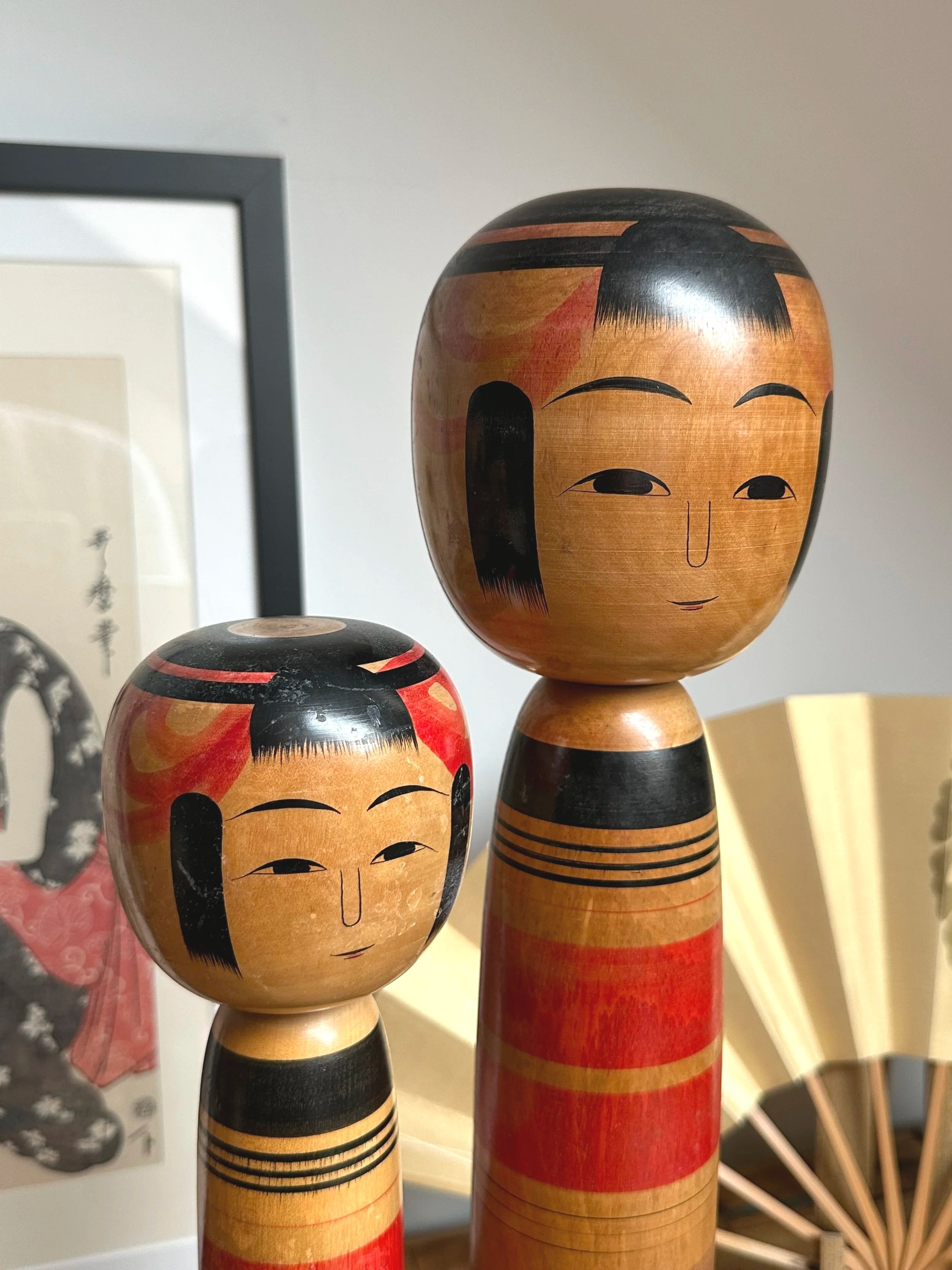 Two Kokeshi dolls by Abe Ichirō, a taller and a shorter doll, with painted wooden bodies banded in red, black, and natural wood, and painted faces.