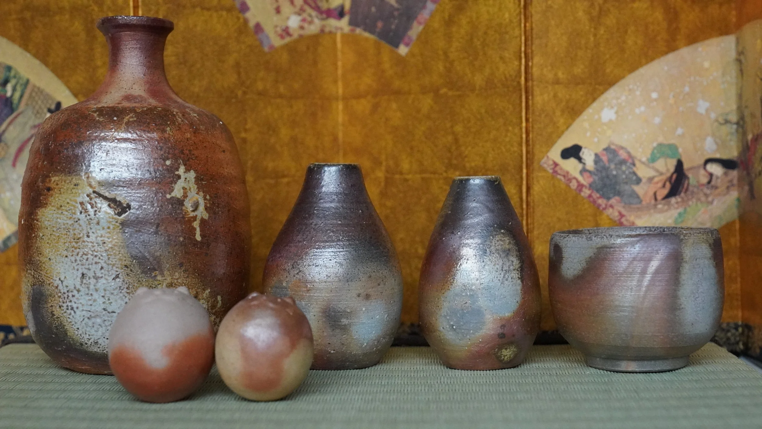 Two textured Bizen-yaki tokkuri vases by Shibaoka Tōsendō, dark aubergine with silver, silvery-blue with rust, on a mat before a golden screen.