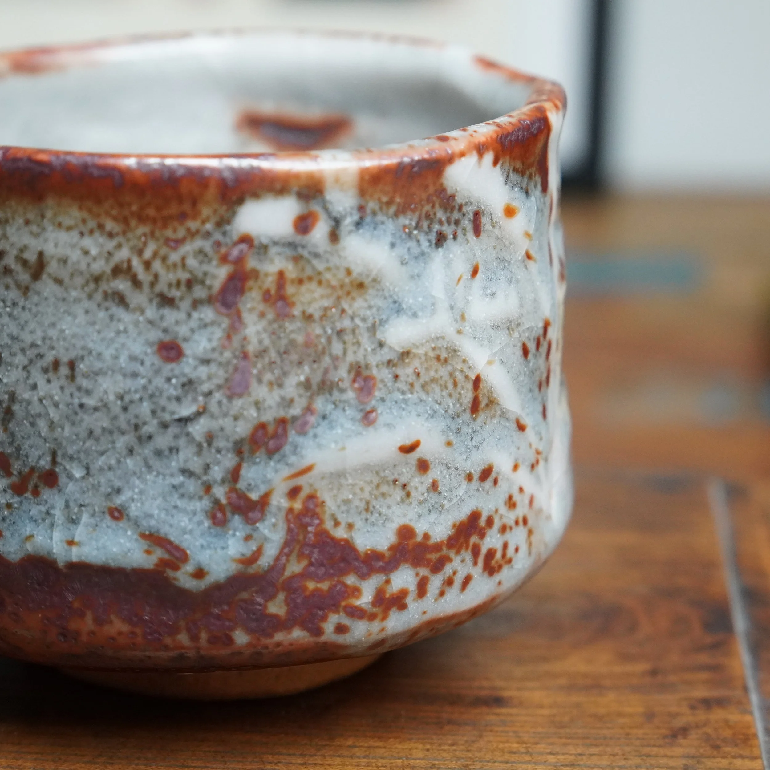 Side view of a winter chawan with its white-grey glaze textured by numerous irregular red-brown speckles, on a wooden surface.