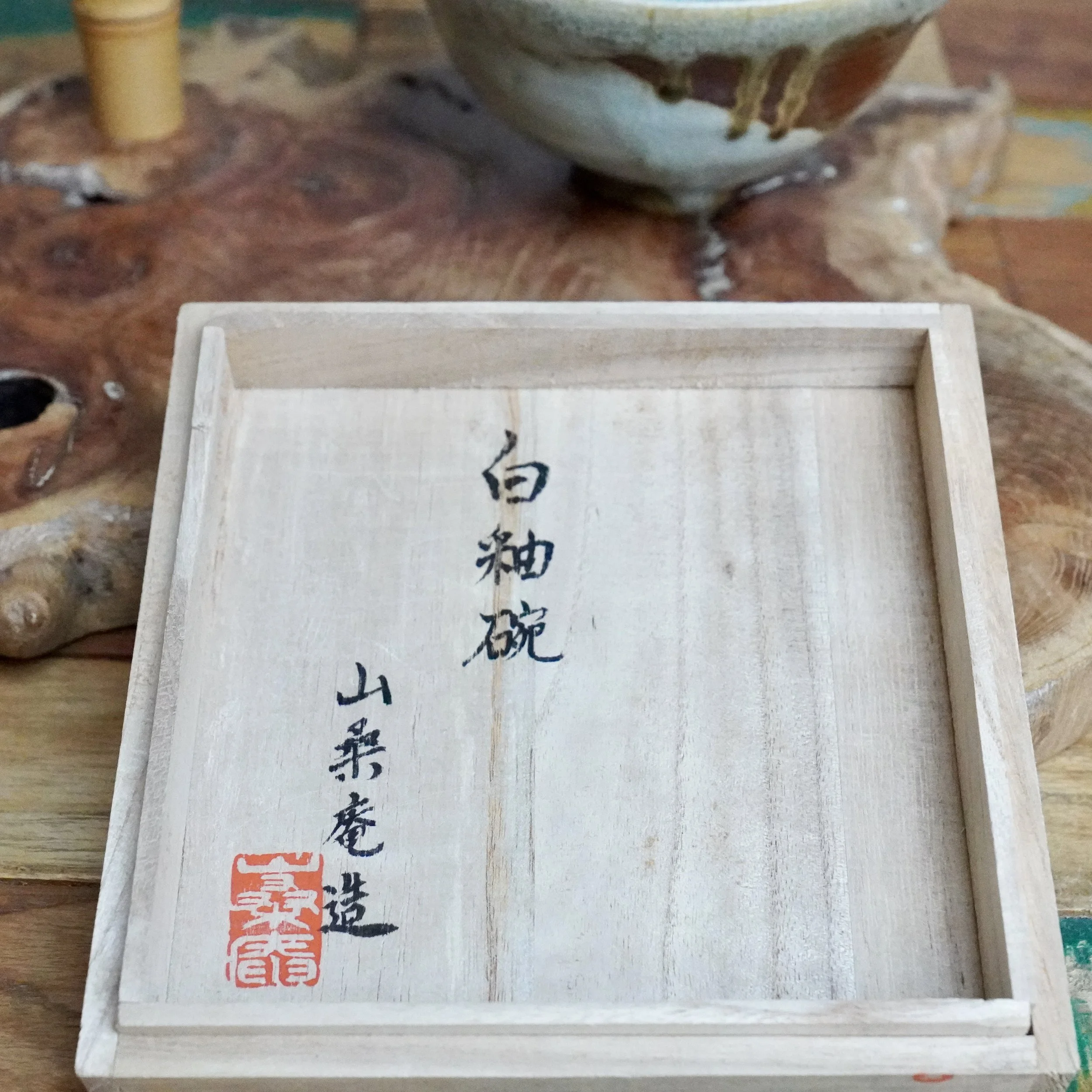 Open wooden tomobako box, a top-down view showing Japanese calligraphy and a red Sansōan seal on its lid. A tea bowl is blurred in the background.