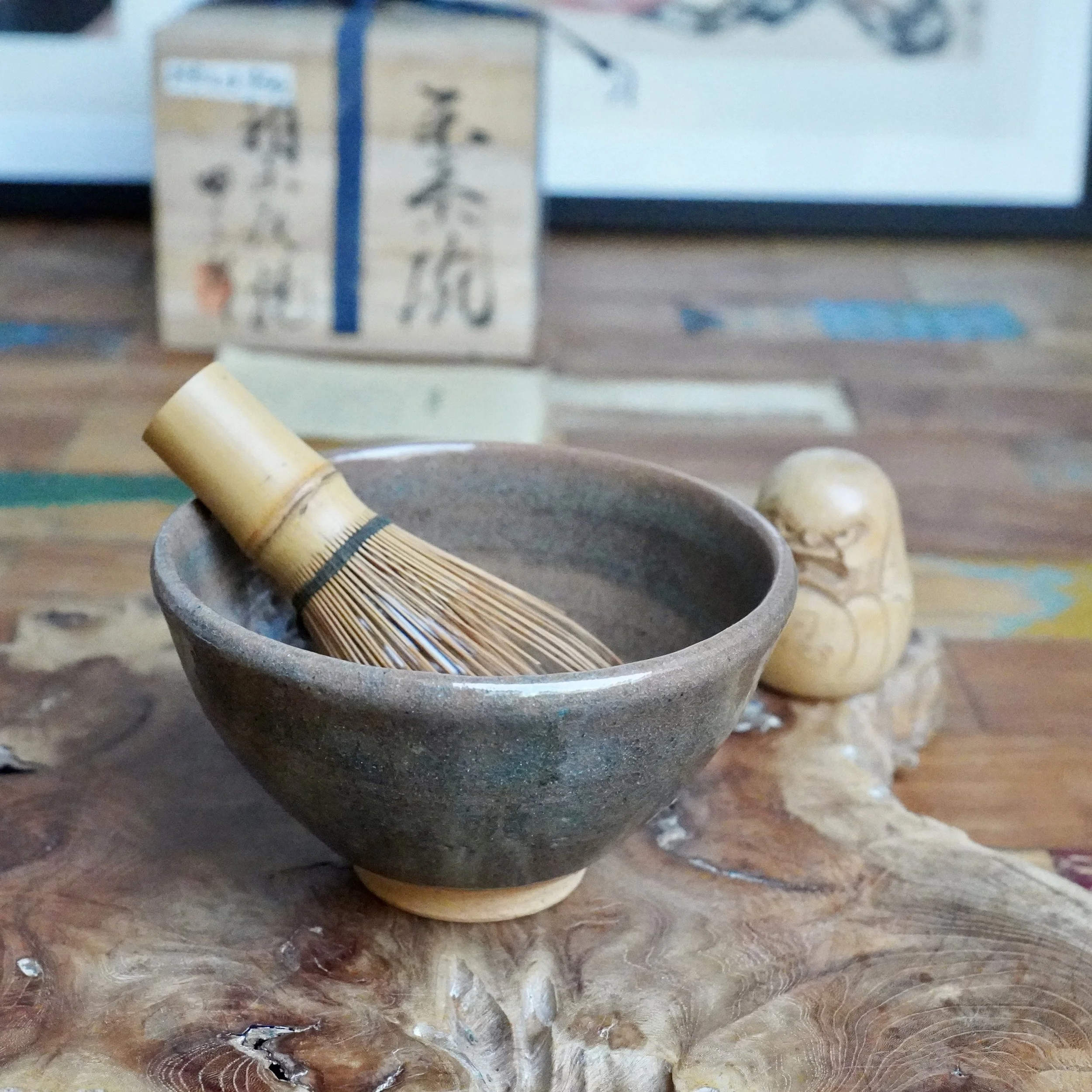 Mizushima Jirō's Kagurao-yaki chawan, dark stoneware with blue-green glaze, bamboo whisk inside, on a wood table with a small figurine and blurred tomobako.