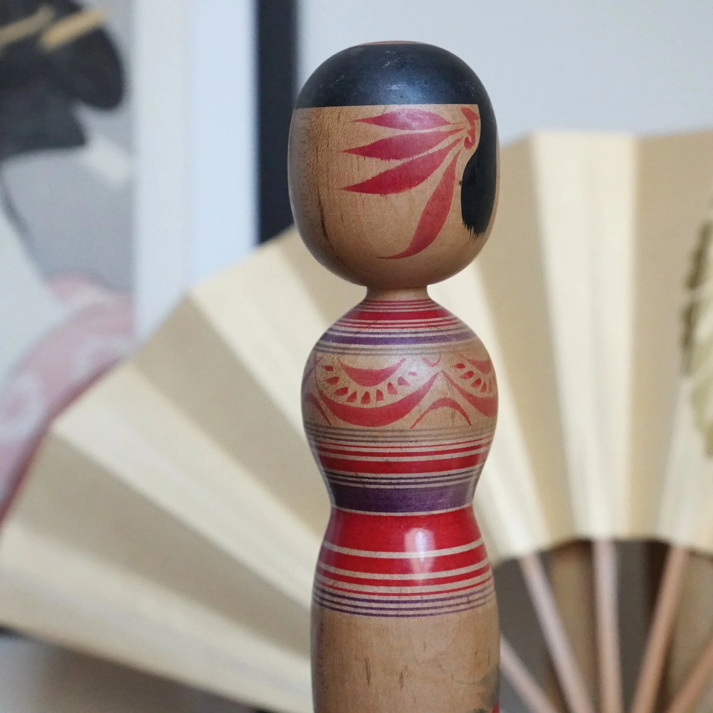 Sasamori Junichi Tsugaru kokeshi: black cap, red leaf patterns on head, red and plum peony motif on wood body with striped waist. Blurred fan in background.