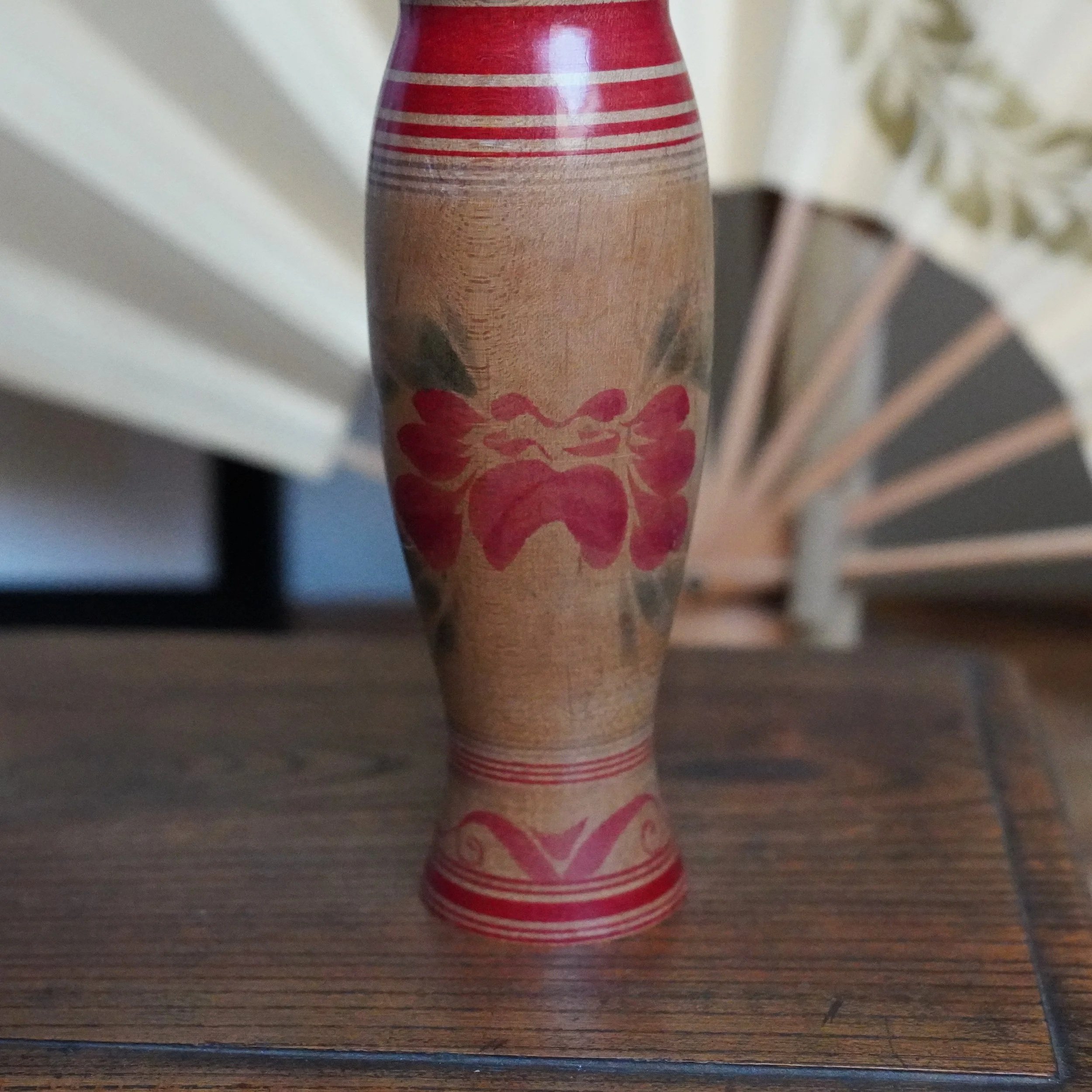 Wooden Tsugaru kokeshi by Sasamori Junichi, with an hourglass shape, red bands, and a red and plum peony motif. On a wooden surface with a blurred white fan.