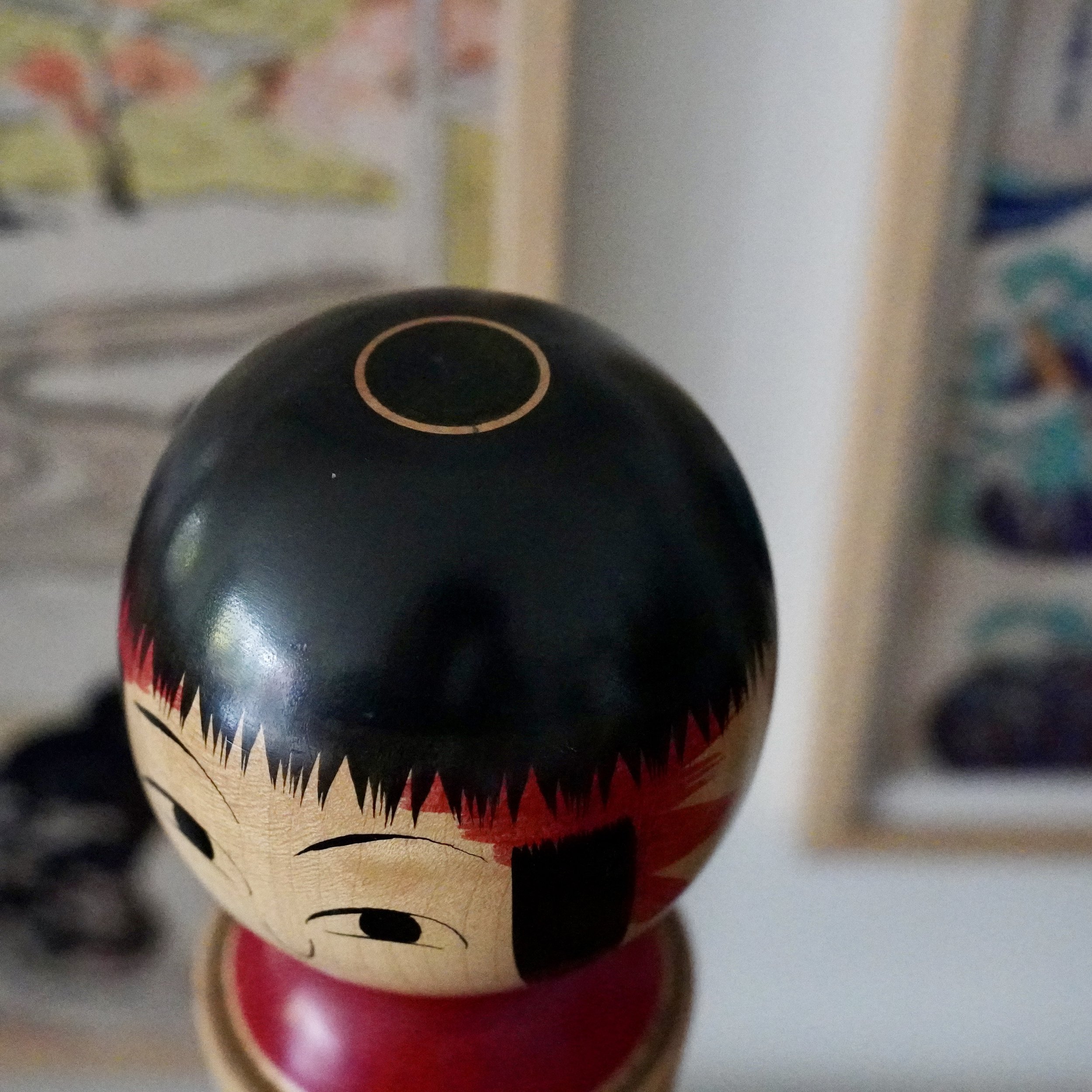 Izu Mamoru Ginzan kokeshi head, glossy black bob, pale wood face with subtle eyes and red collar. A thin golden circle decorates its crown.