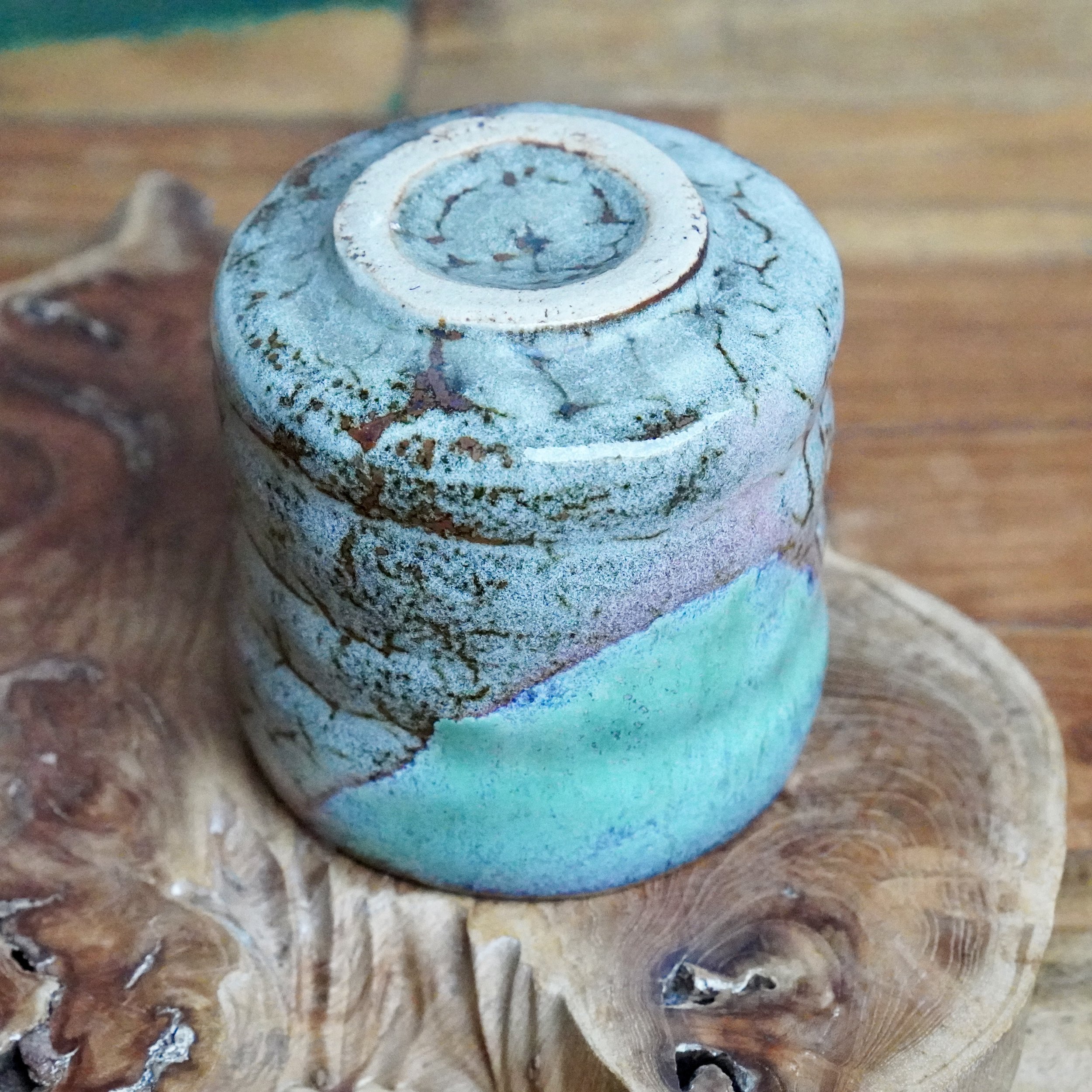 Hand-thrown tsutsu chawan, underside up. Speckled blue-grey glaze with brown lines, a clear turquoise band. Unglazed base rests on a wood slice.