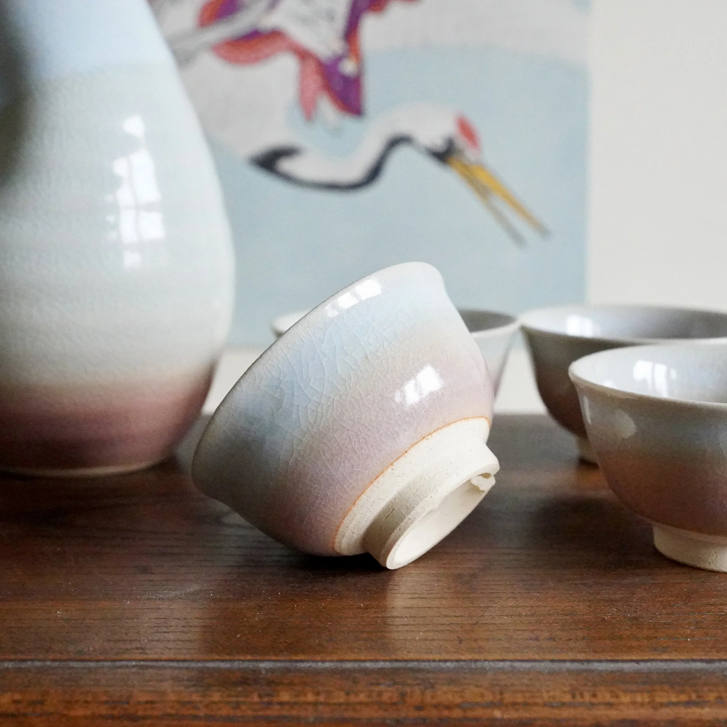 Hagi-yaki sake set: a tokkuri and four ochoko with blush to pale blue-grey crackle glaze. One cup is tilted on a wood surface, with a crane artwork behind.