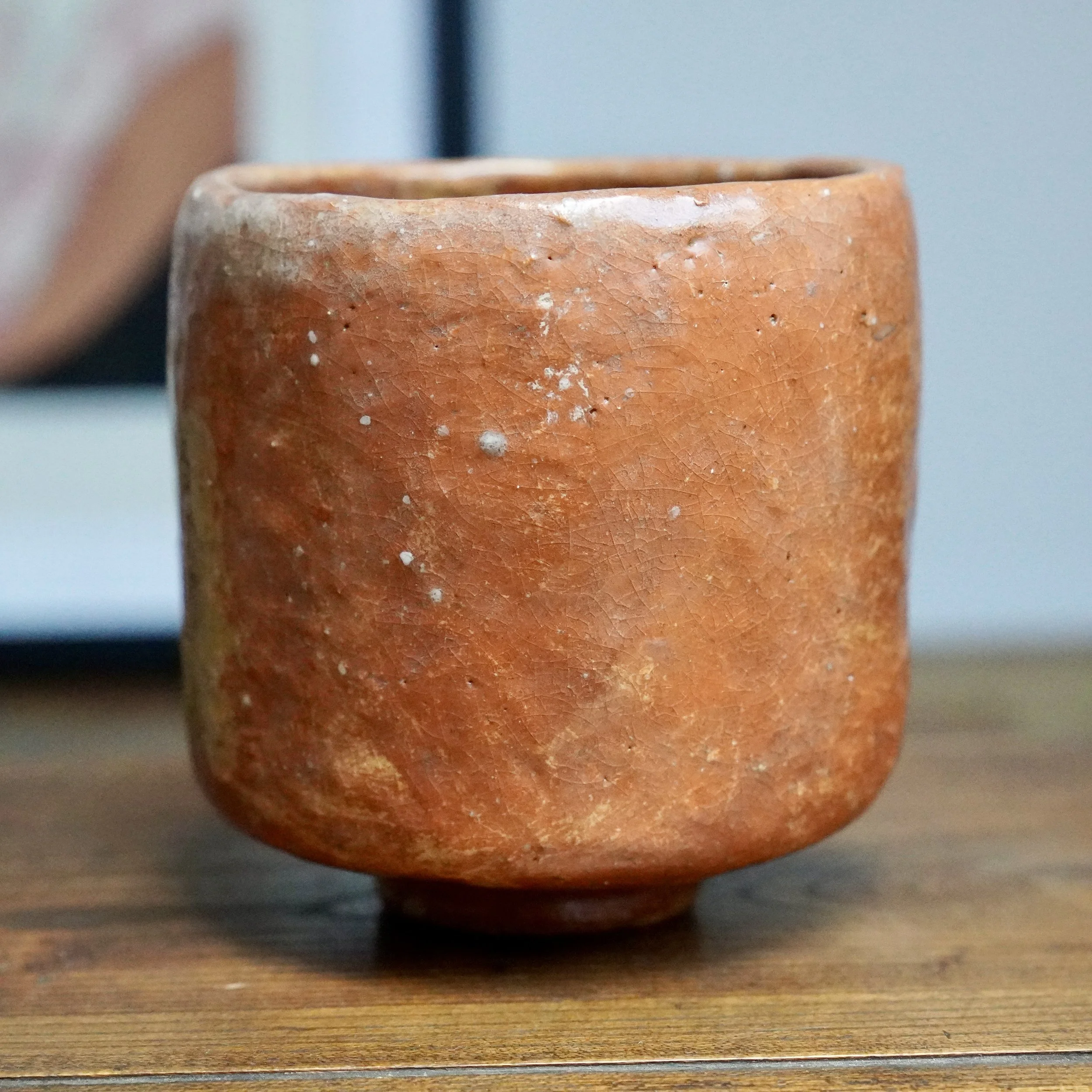 Reddish-orange tsutsu chawan by Hioki Nanshū kiln, with a crazed texture and irregular white glaze marks, on a wooden table.