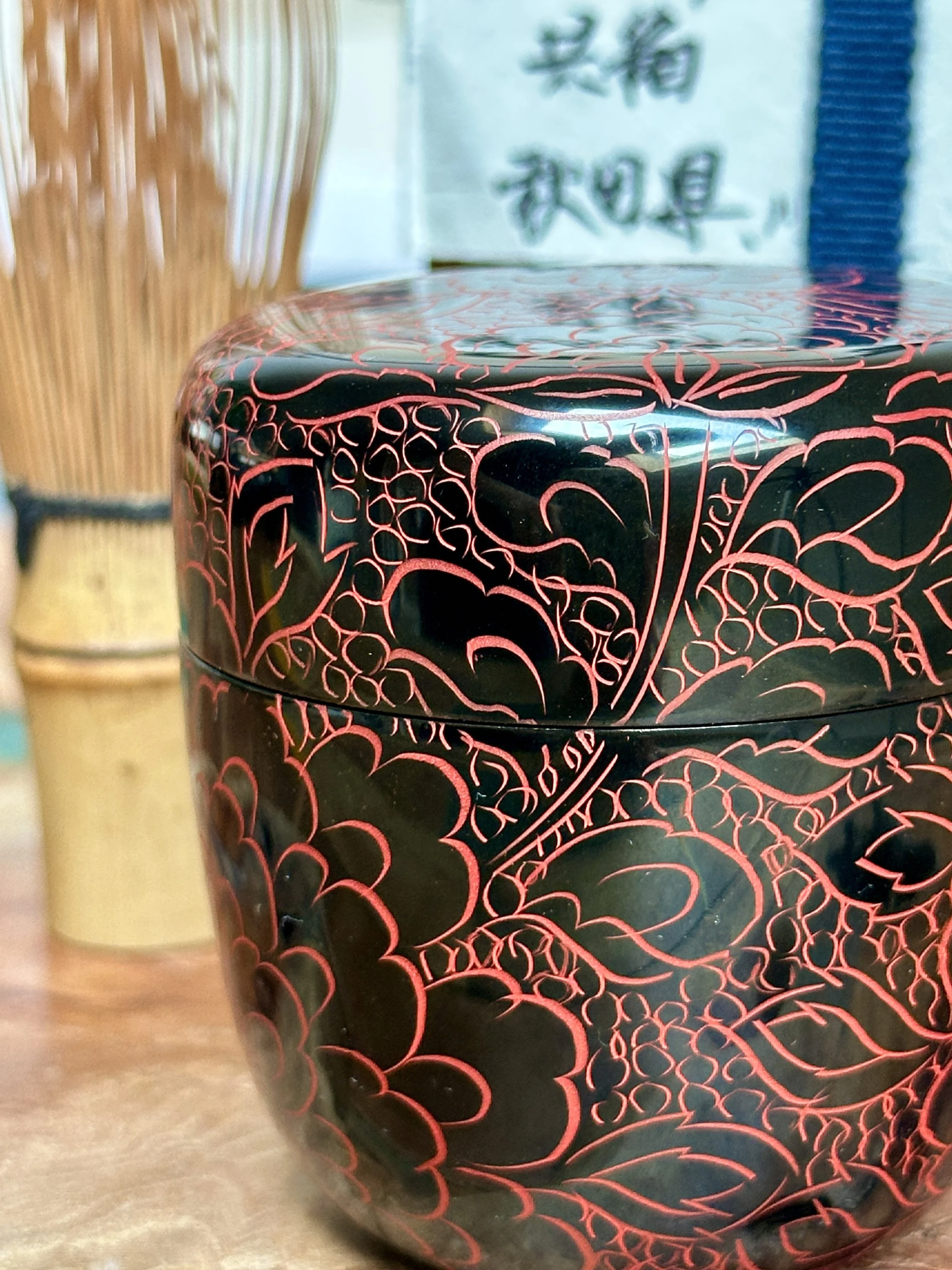 Rounded, glossy black lacquered tea caddy, featuring intricate vermilion peony designs engraved across the lid and body. A blurred bamboo whisk is visible left.