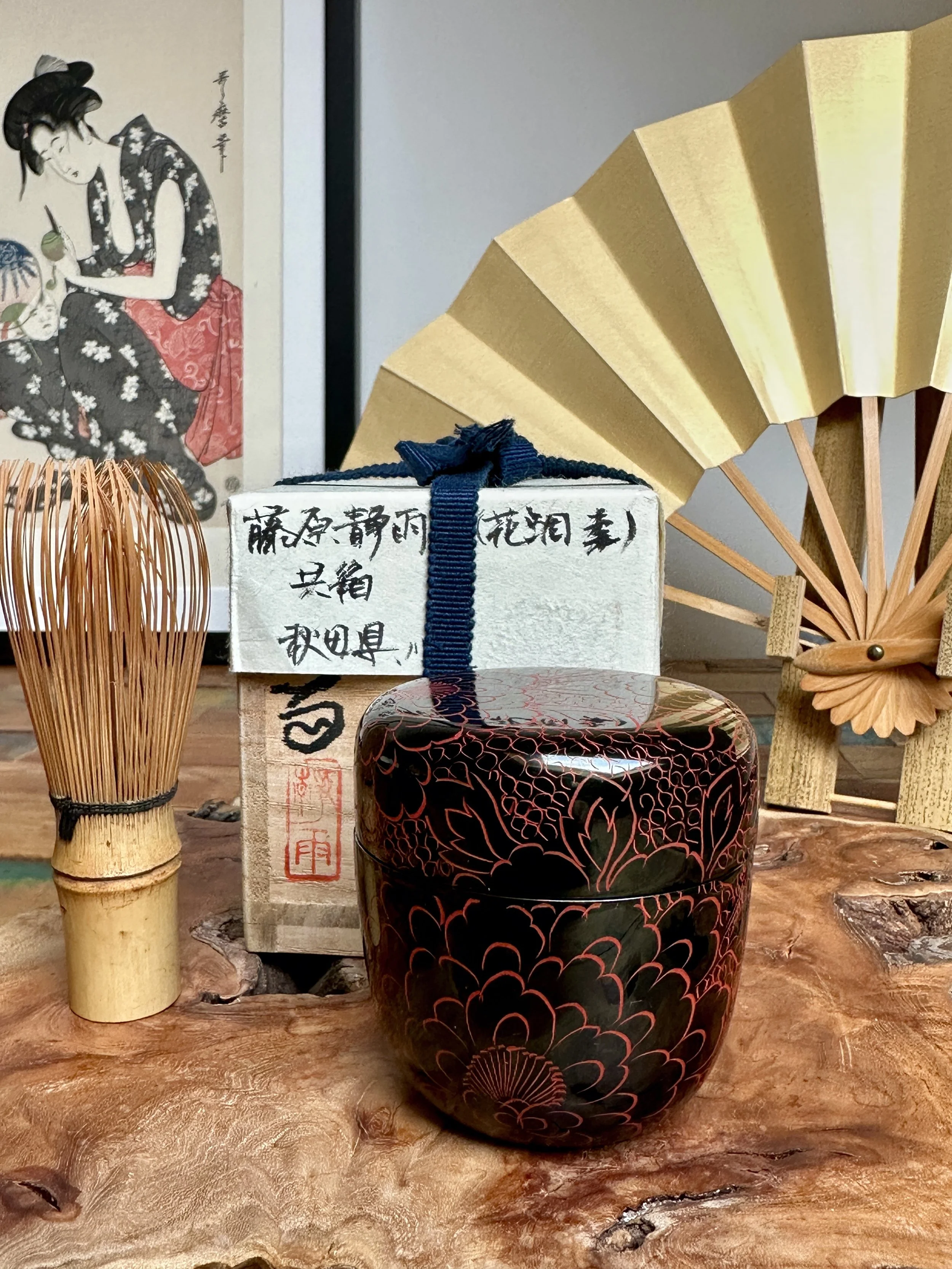 Black lacquered tea caddy with red peony designs, a bamboo whisk, and a box signed Fujiwara Seiu, on a wooden surface with a fan and print.