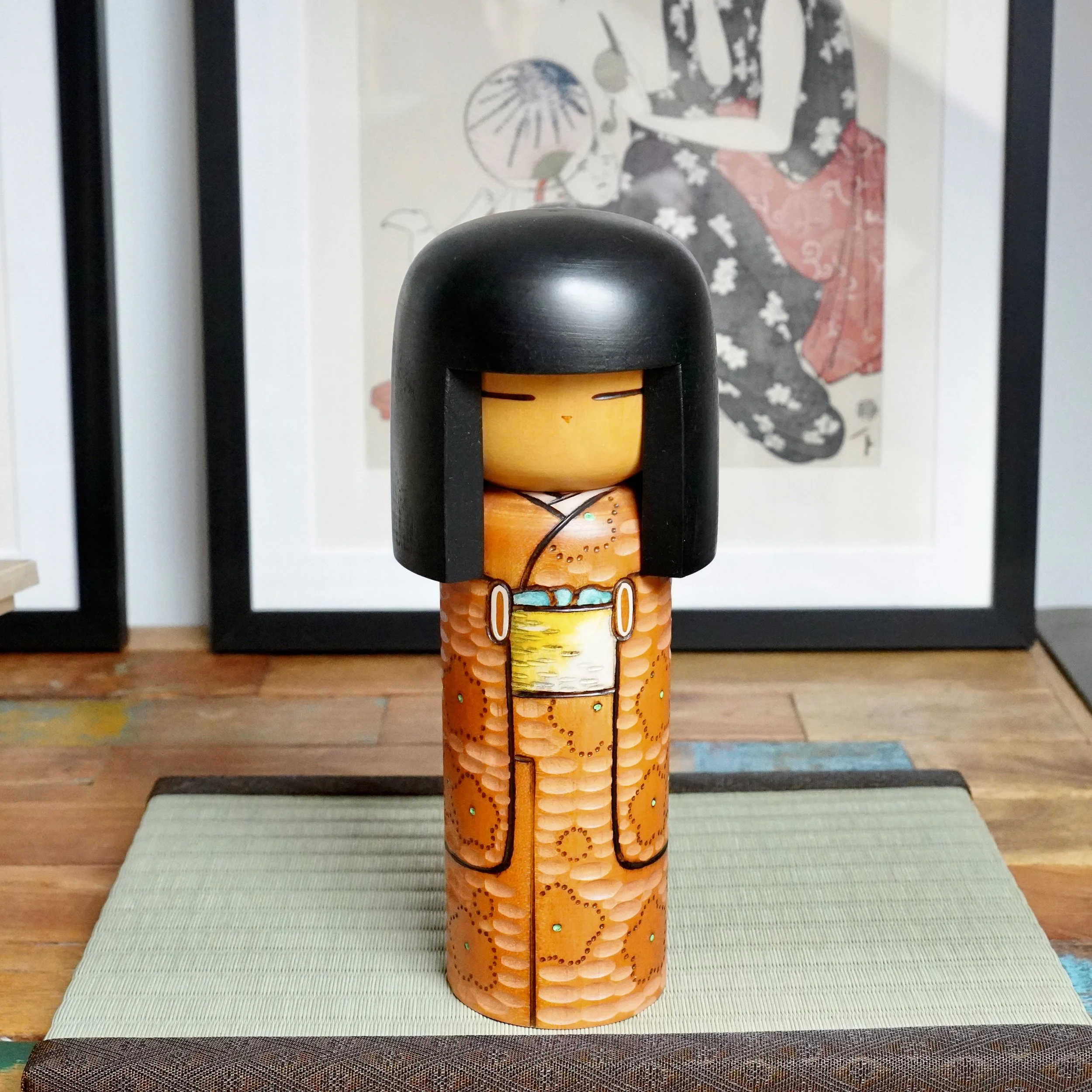 Front view of a Kisaku sōsaku kokeshi doll with black hair, an orange carved kimono detailed with green dots, and a yellow obi, on a green woven mat.