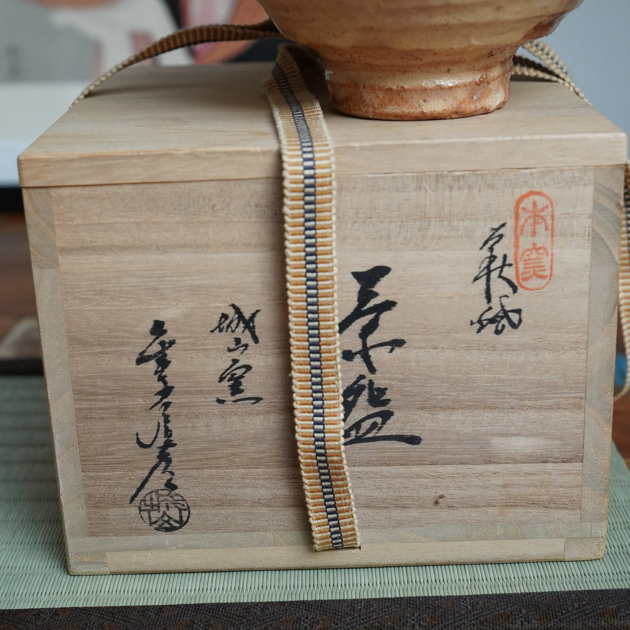 Wooden tomobako, signed by Kaneko Nobuhiko with black calligraphy and a red seal, tied with a striped cord, with a ceramic chawan resting on top on a mat.